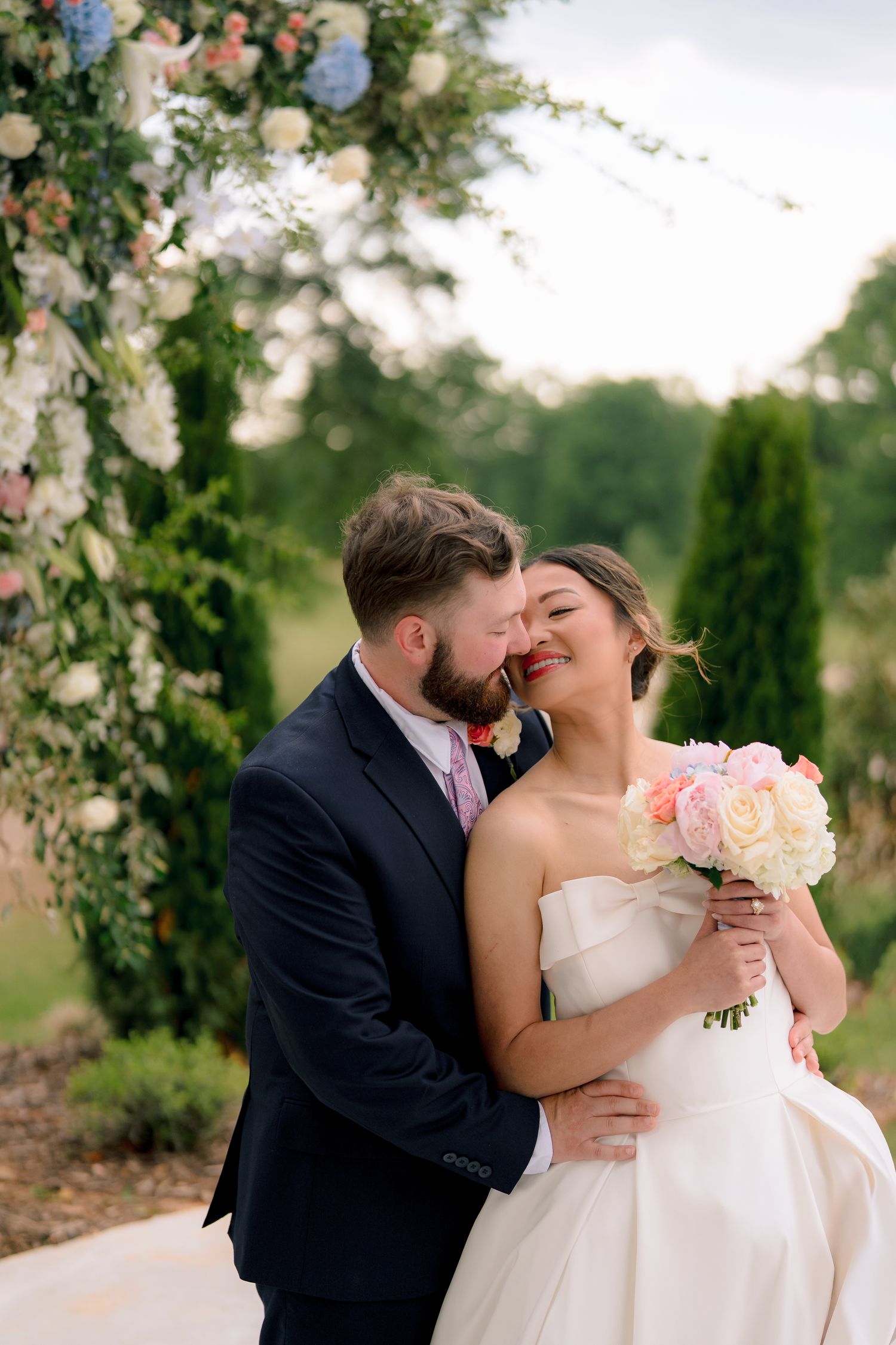 Bride and groom share a romantic kiss under a floral arch outdoors, bride holding a pink and white bouquet.
