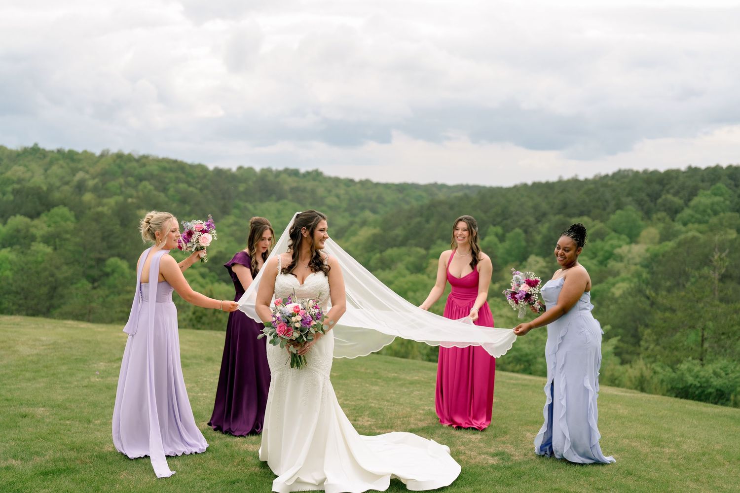 Bride in white gown with bridesmaids in colorful dresses holding her veil on a scenic hilltop.