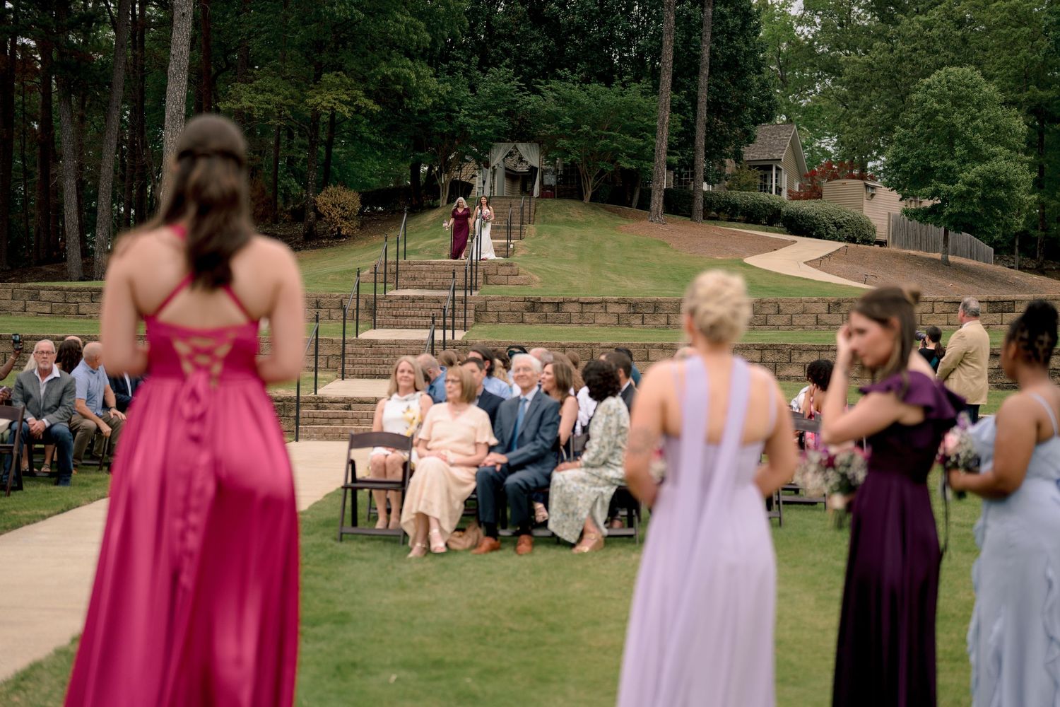 Bridesmaids in colorful gowns watch as guests gather for an outdoor wedding ceremony on a lush green hillside.