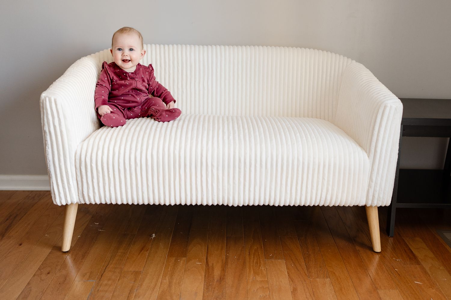 Small baby wearing burgundy outfit sits on modern white ribbed loveseat with wooden legs.