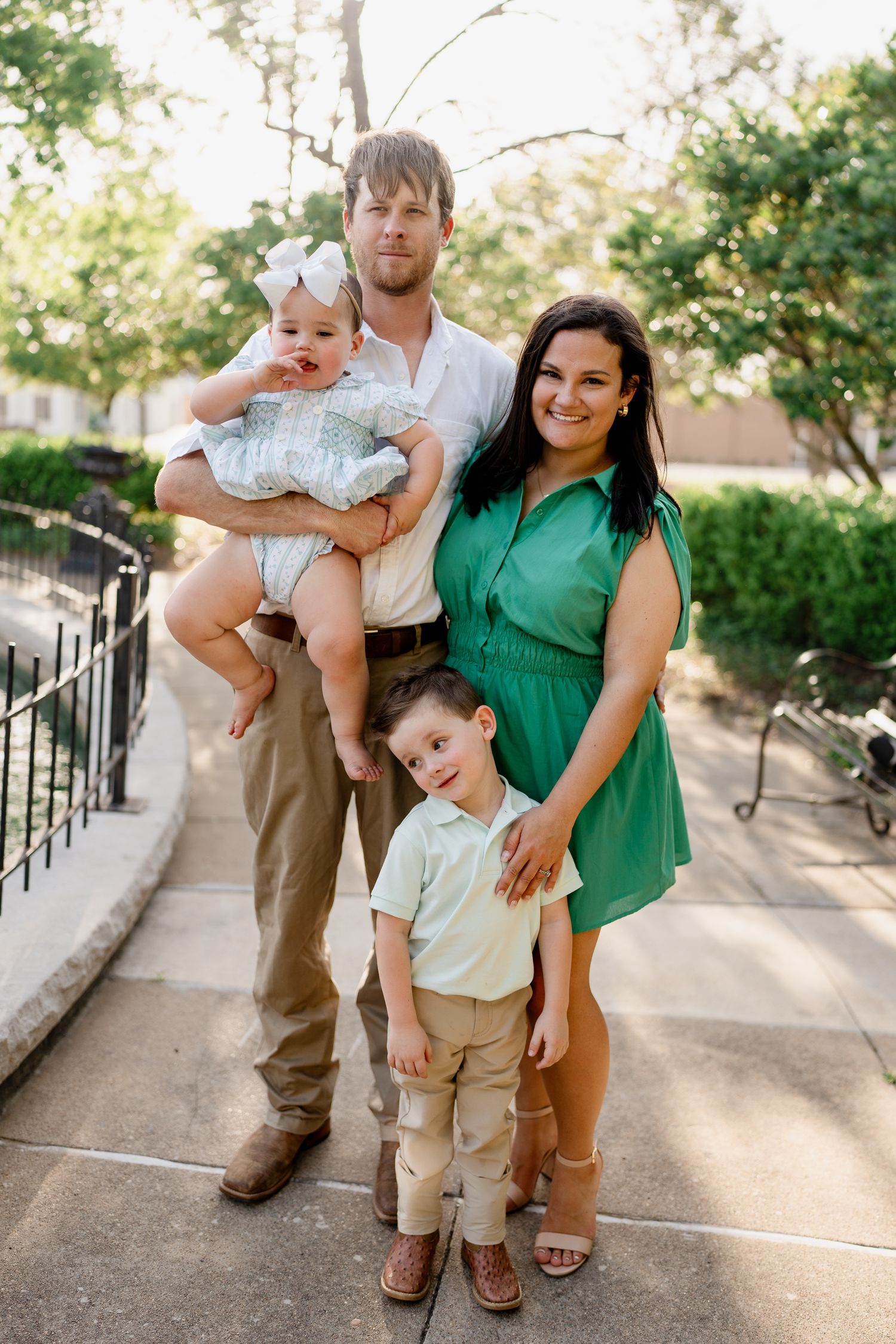 Family poses playfully on concrete path surrounded by greenery in coordinated green and khaki outfits.