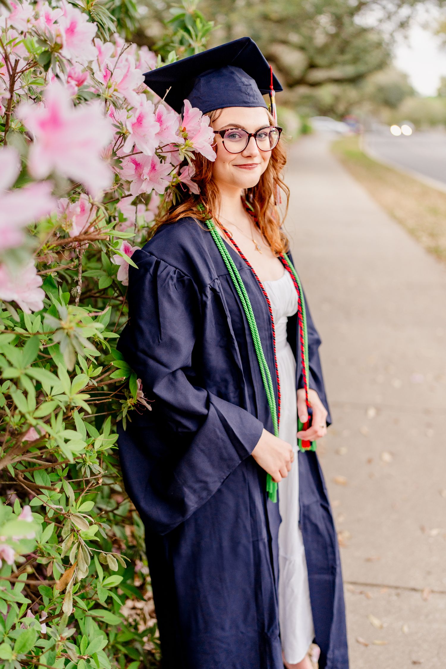 Graduate in navy blue academic regalia poses beside pink flowering bushes.