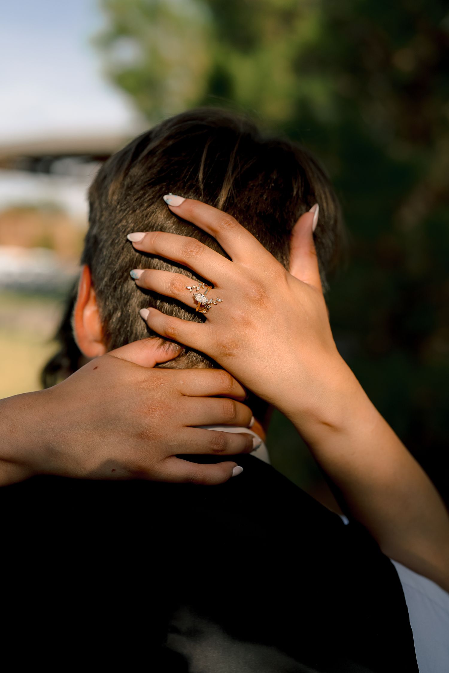 Close-up of bride's hand with gold diamond engagement ring resting on groom's head during outdoor wedding.