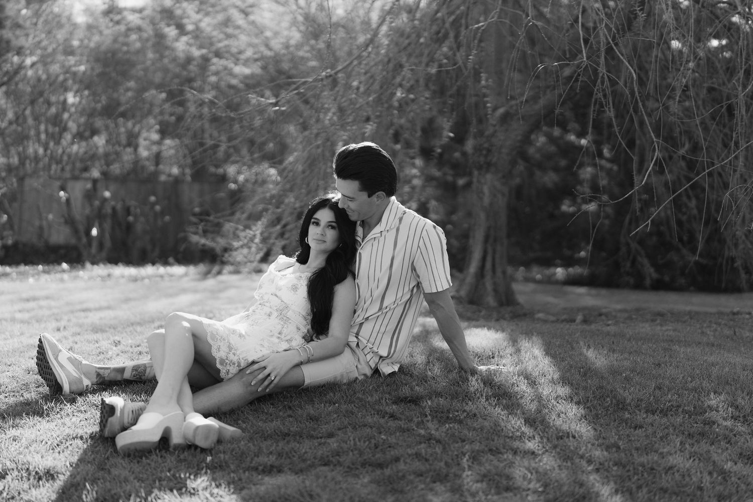 Black and white photo of a couple relaxing together on grass beneath a large willow tree.