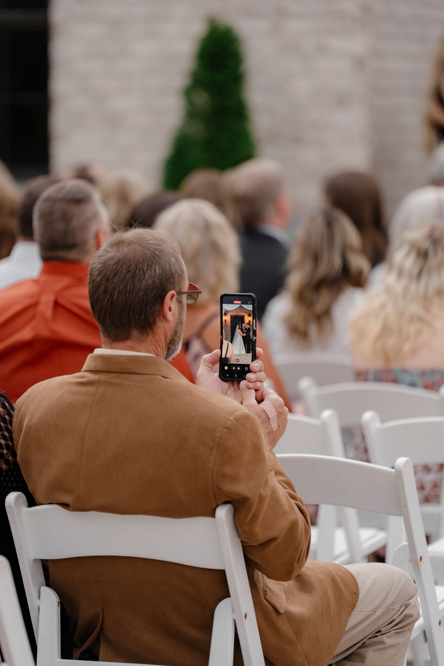 Wedding guest in brown jacket captures photos on phone during outdoor ceremony.