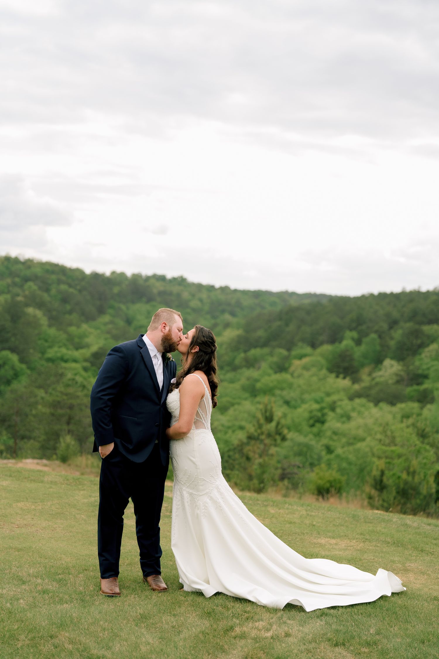Bride and groom share a romantic kiss on a green hillside, with lush forest backdrop on their wedding day.