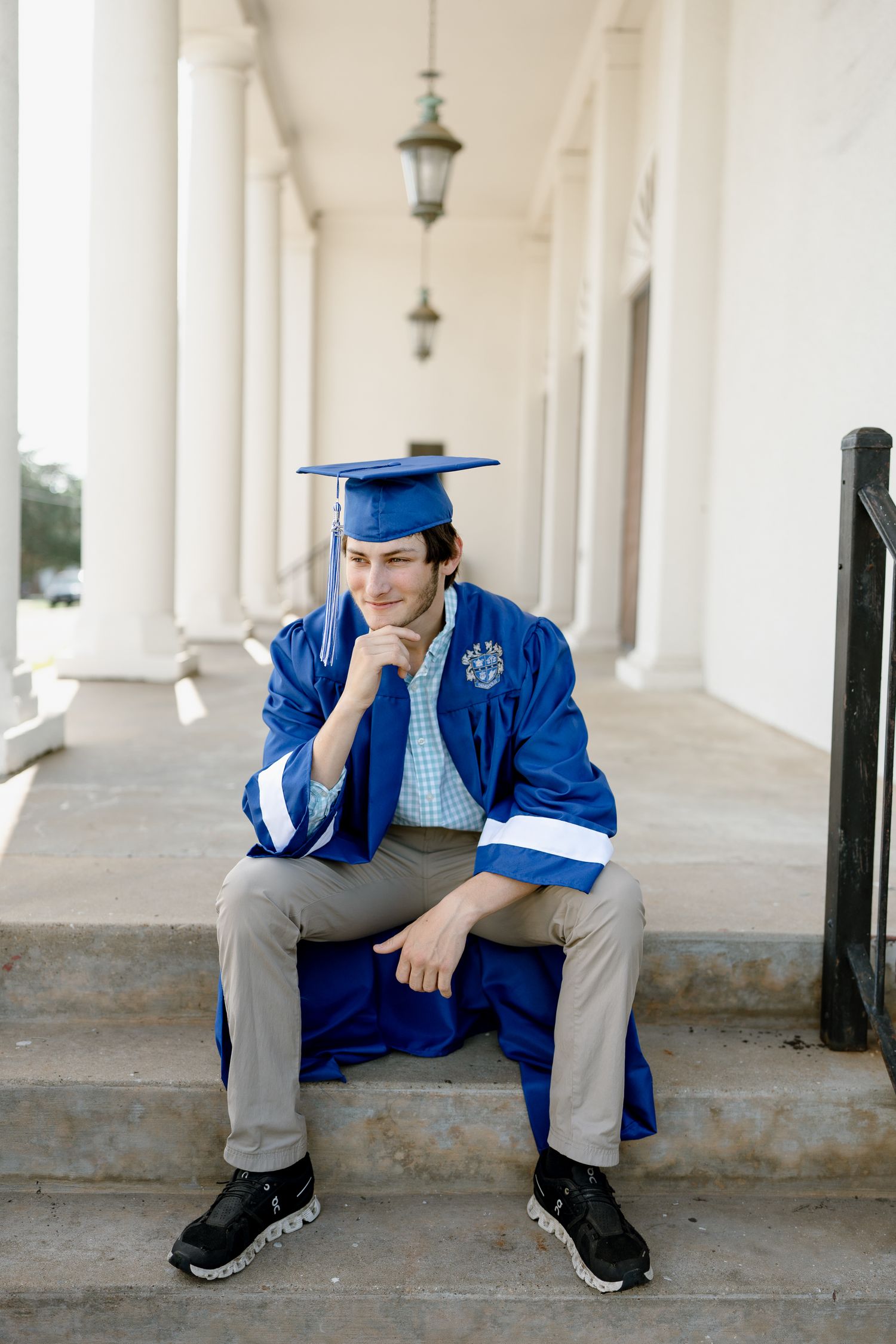 Graduate in blue regalia sits thoughtfully on steps of columned building.