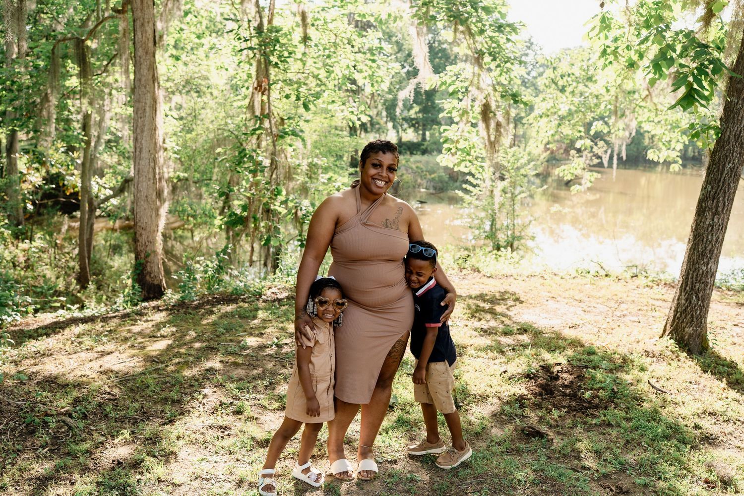Woman in beige dress and child enjoying a sunny day by a river in a wooded setting.