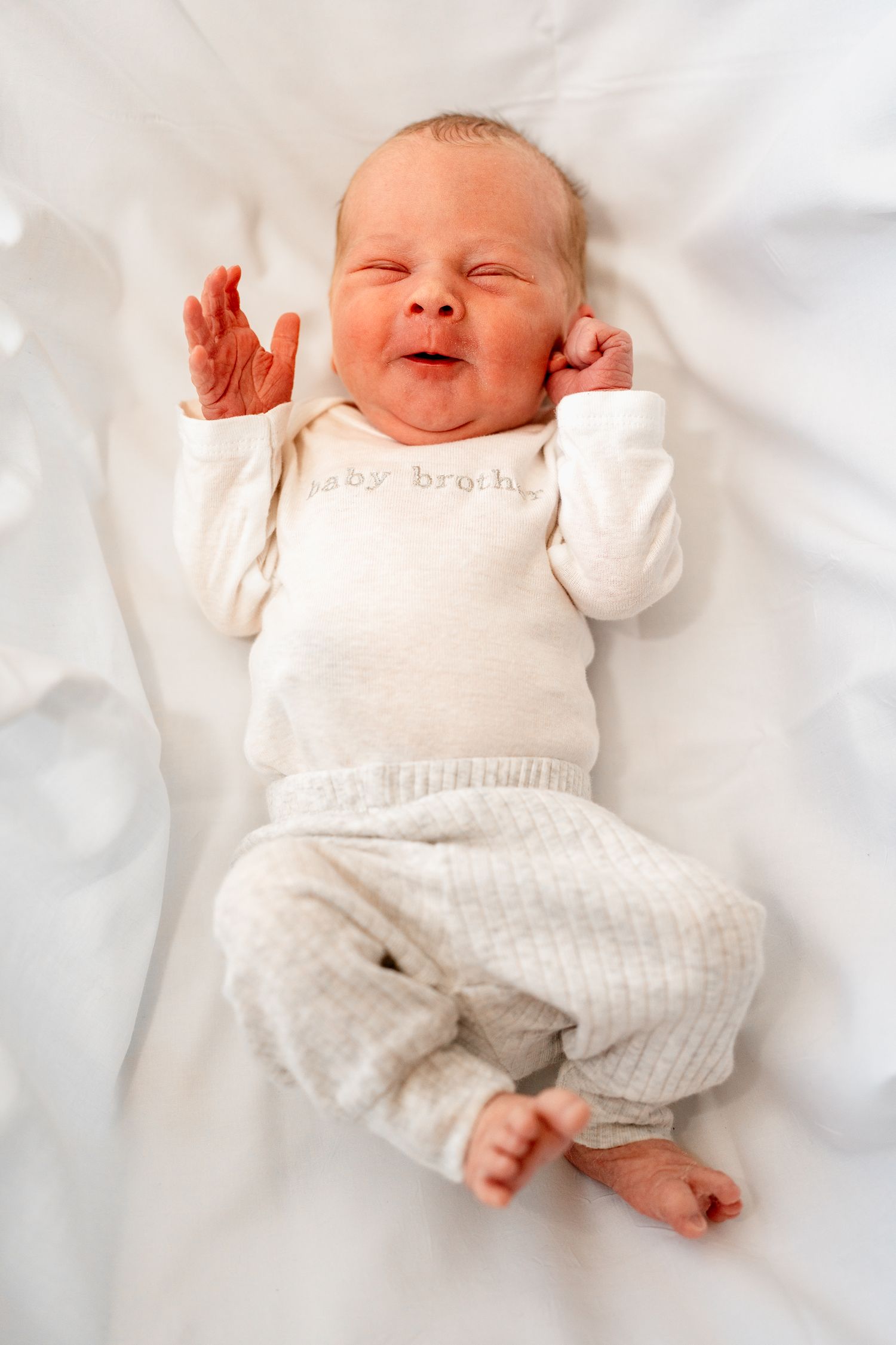 Newborn in cozy white outfit stretches while resting on clean white sheets.
