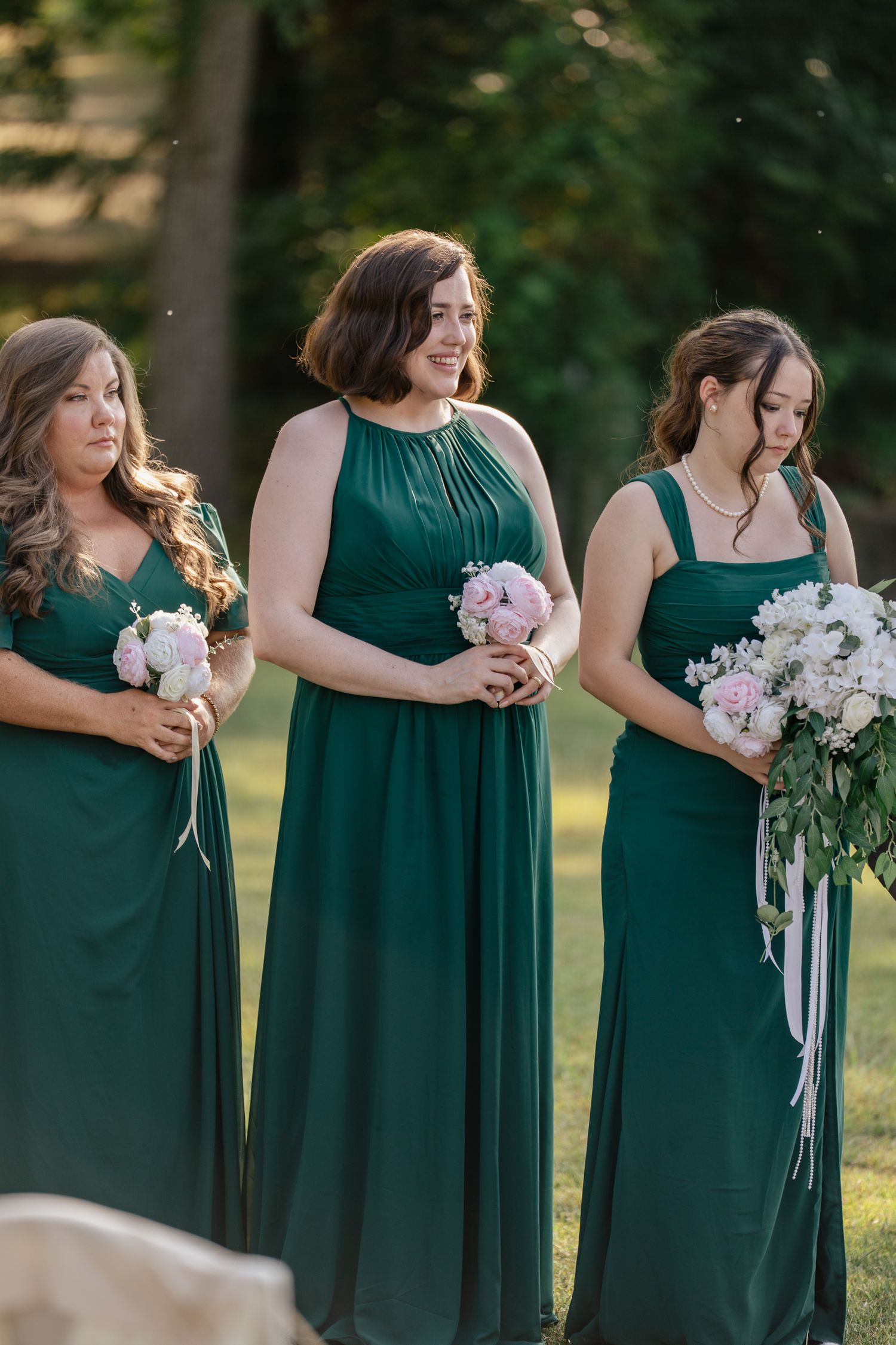 Three bridesmaids in emerald green floor-length dresses hold pink and white bouquets during an outdoor wedding ceremony.