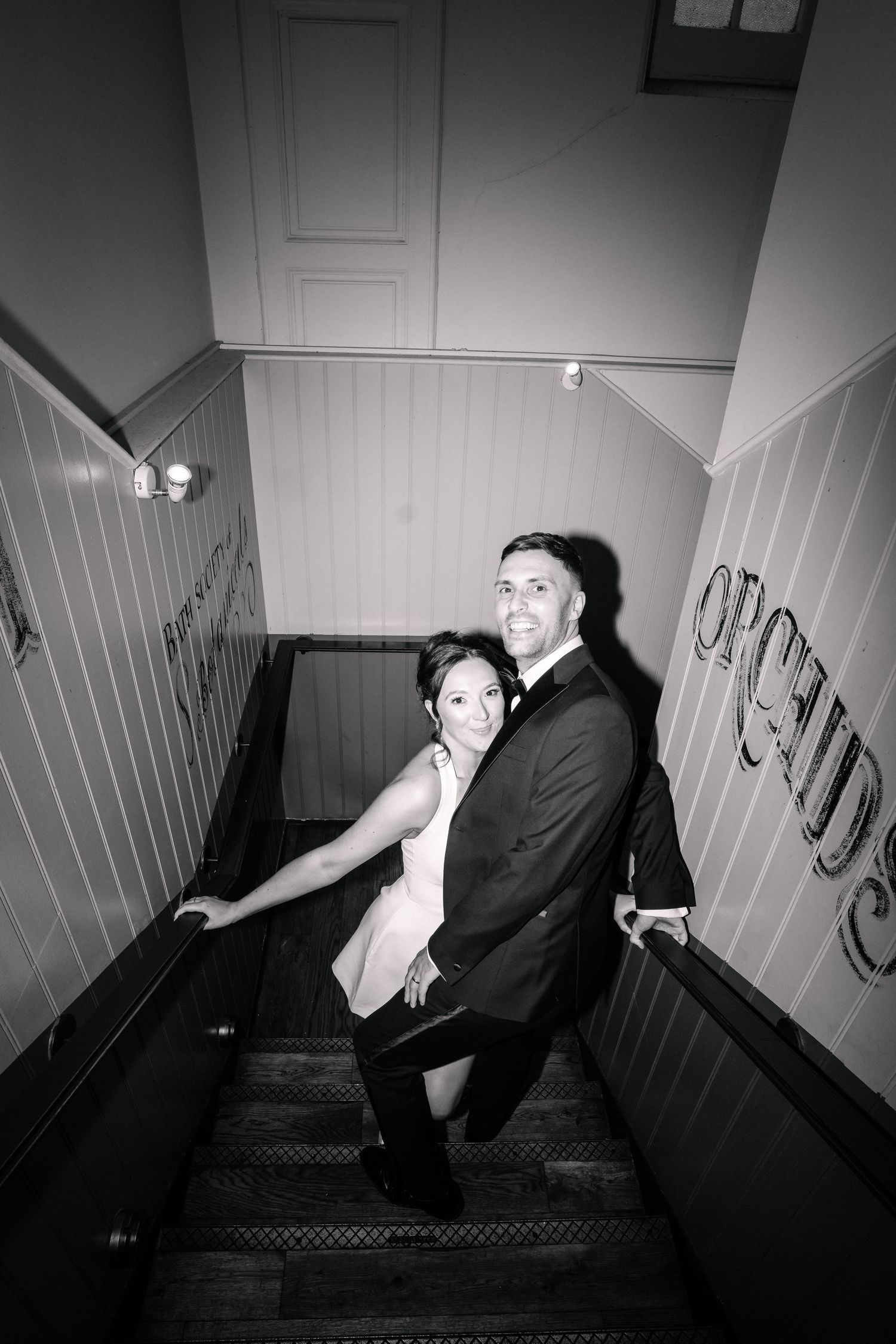 Black and white photo of a couple posing on stairs, man in tuxedo and woman in dress, smiling at camera.