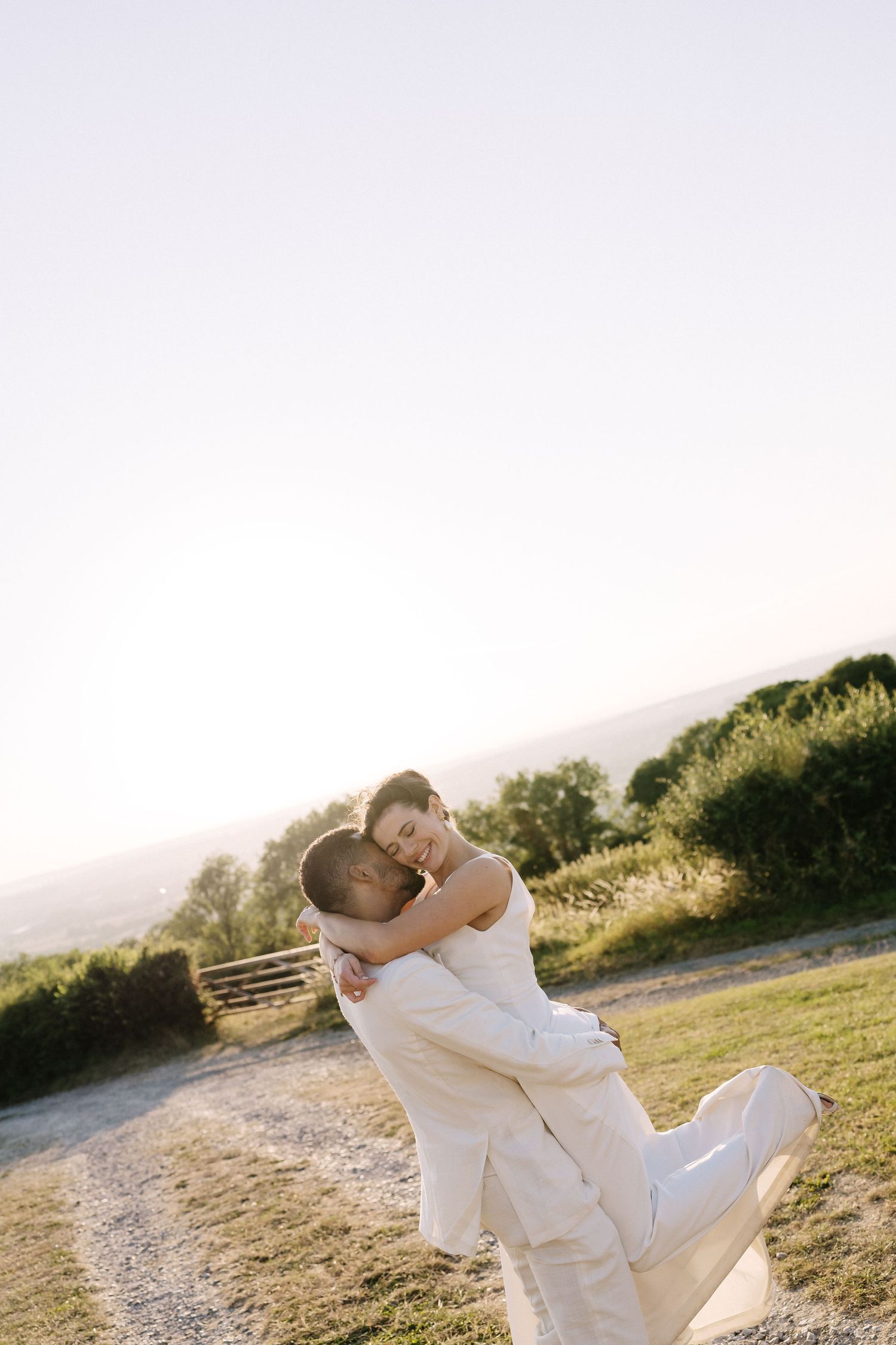Couple in white attire sharing a romantic embrace outdoors at golden hour with scenic countryside backdrop.
