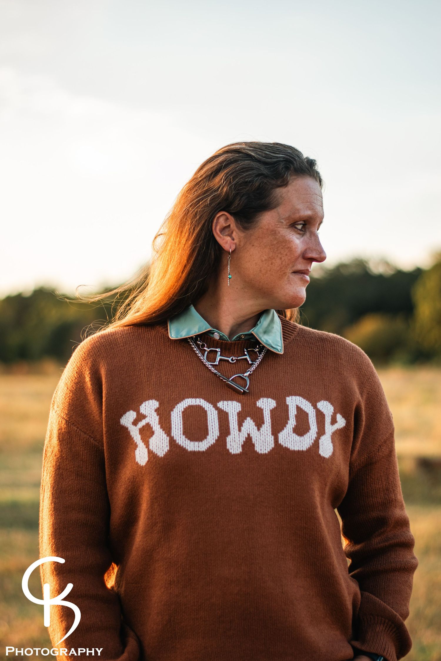 Rustic outdoor portrait photography featuring a woman in a “Howdy” sweater, captured in golden hour light with warm country atmosphere.