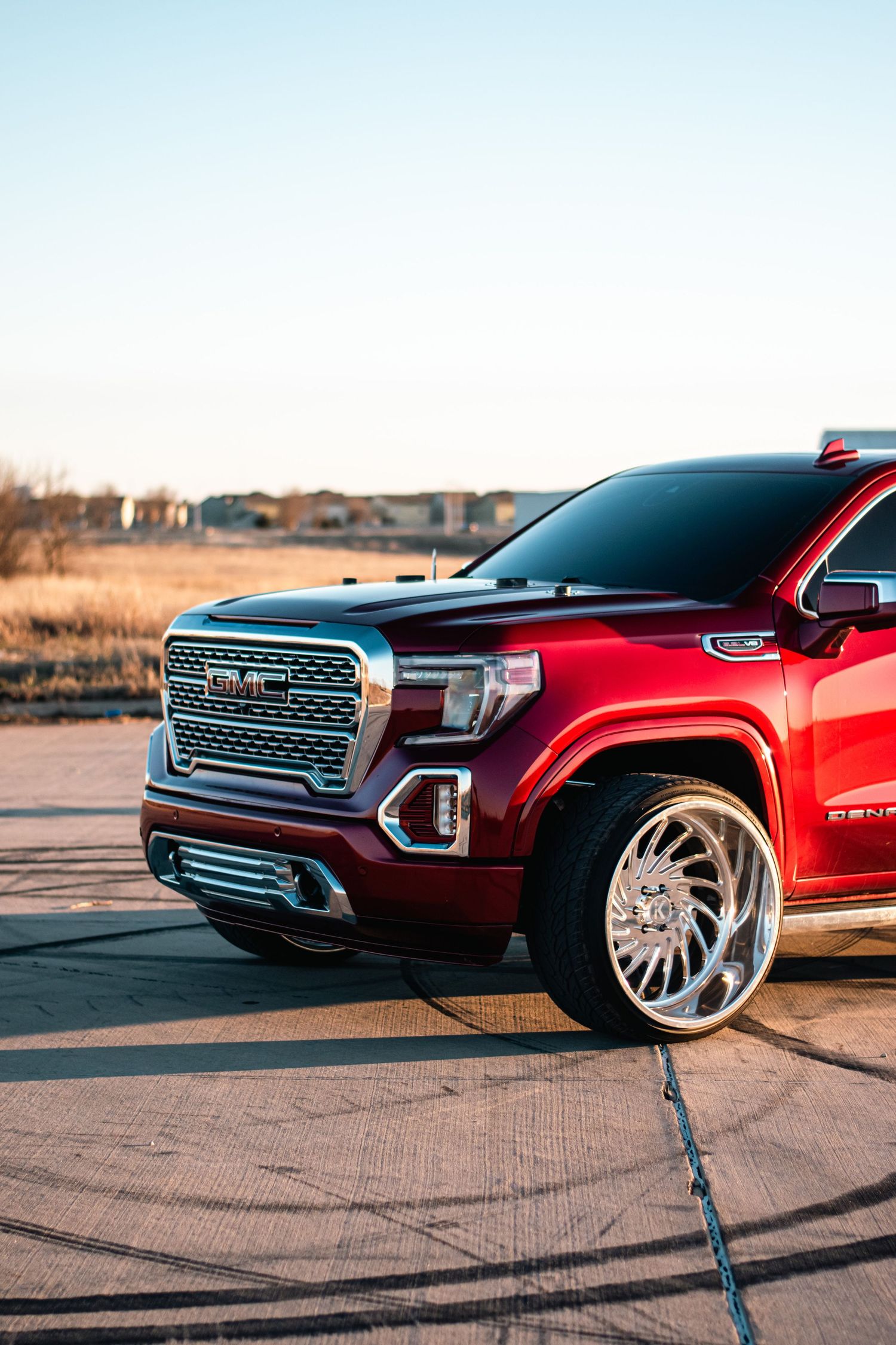 Close-up automotive photography of a custom red GMC Sierra, highlighting chrome accents, polished wheels, and bold styling in golden light.