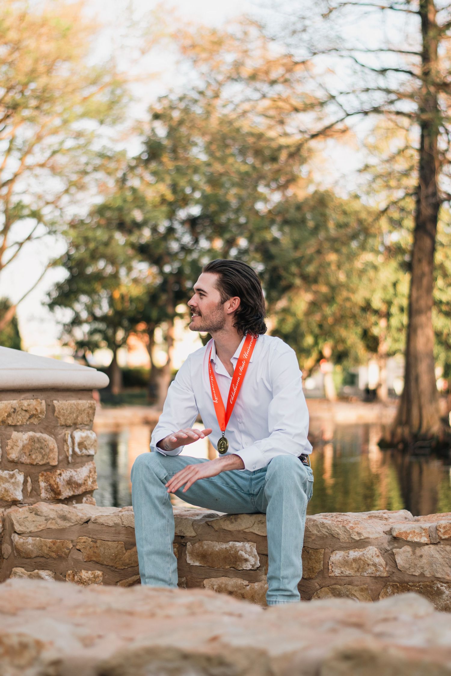 Outdoor graduation portrait photography of a young man with medal, seated by stonework and water, framed by trees in warm natural light.