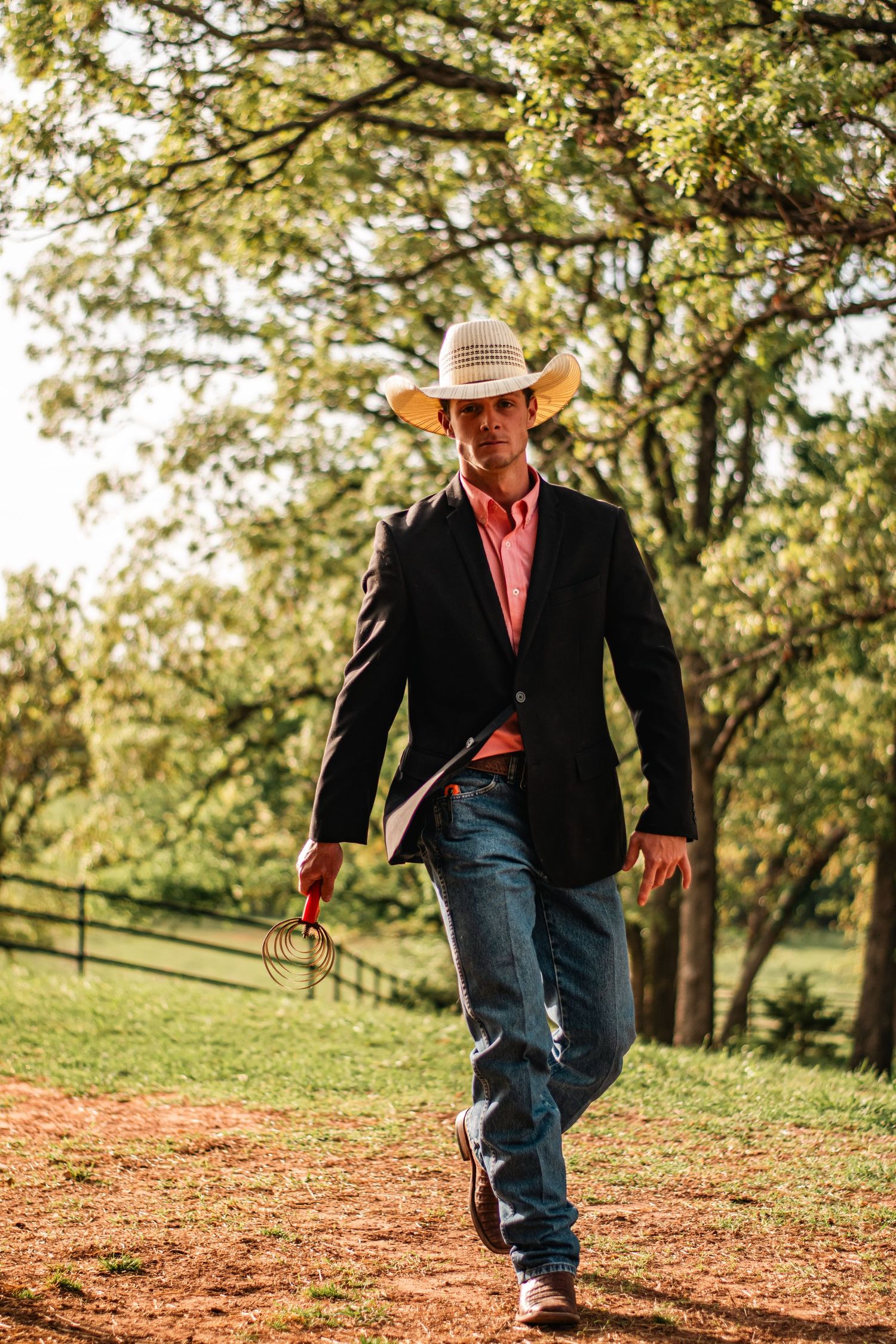 Western portrait photography of a young man in cowboy hat, jeans, and blazer walking outdoors, captured in warm natural evening light.