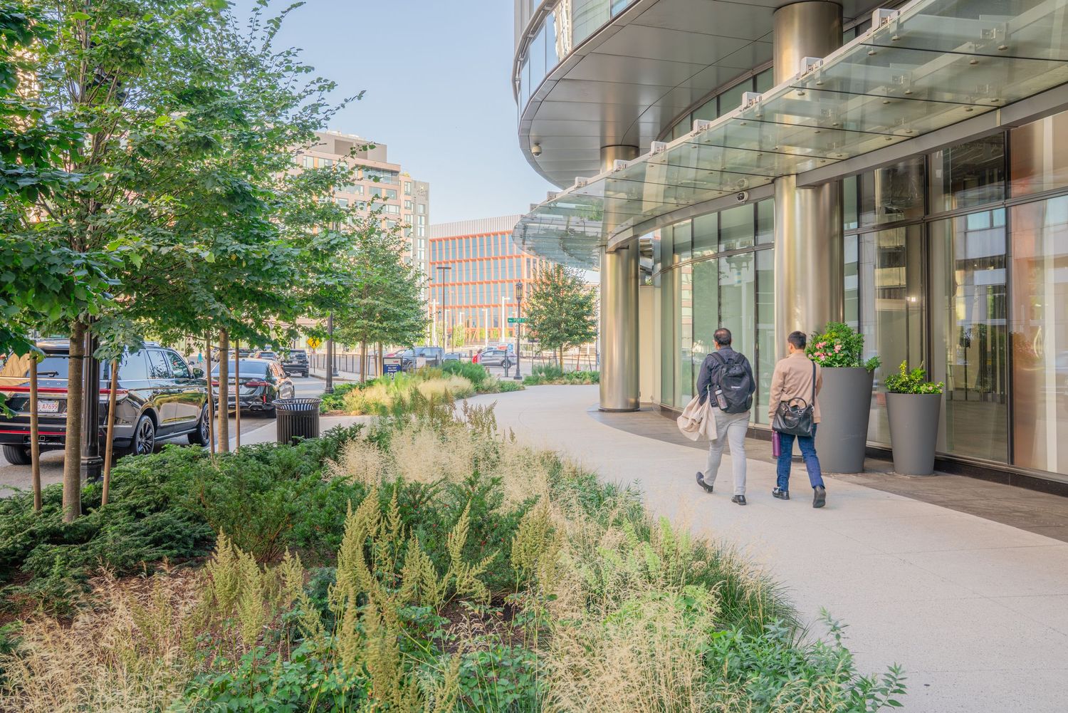 Contemporary walkway along glass-fronted building with integrated landscaping and seating areas.