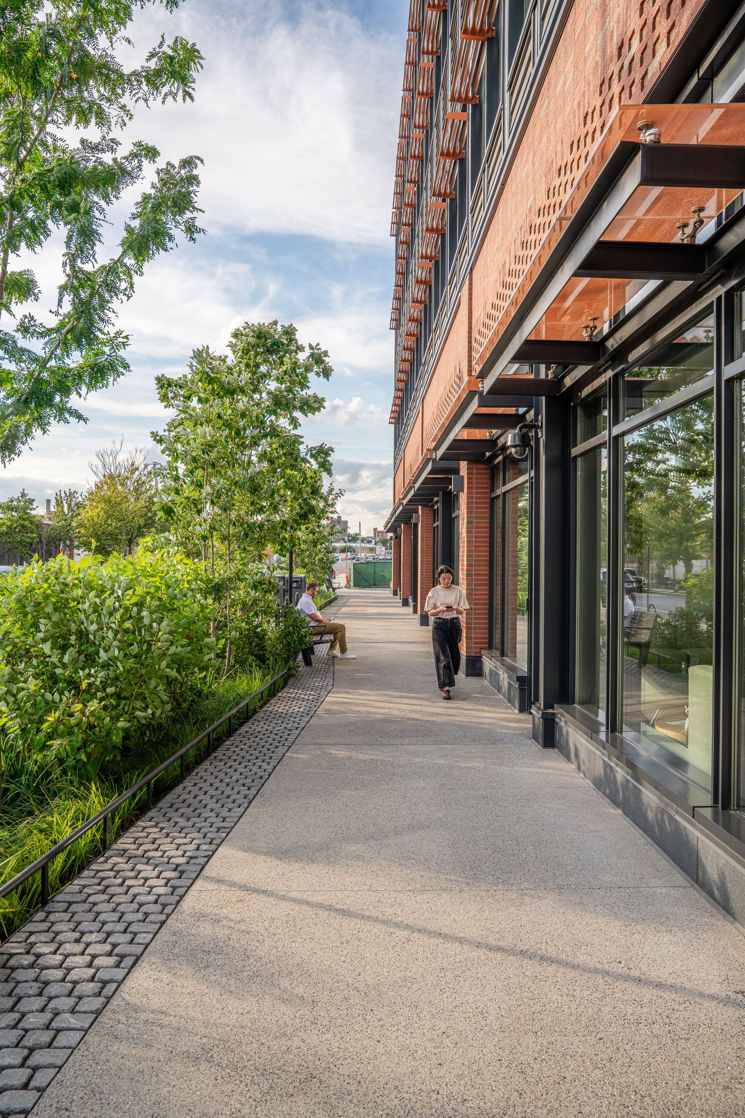 Wide concrete walkway alongside brick building with architectural overhangs and green landscaping.