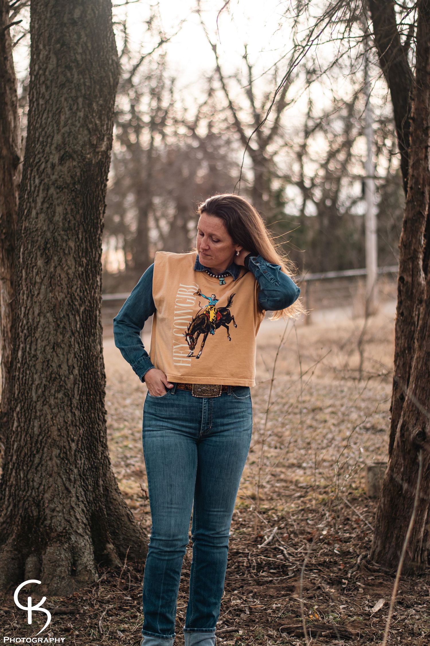 Western lifestyle portrait photography of a woman in denim and graphic vest, captured outdoors among trees in soft natural evening light.