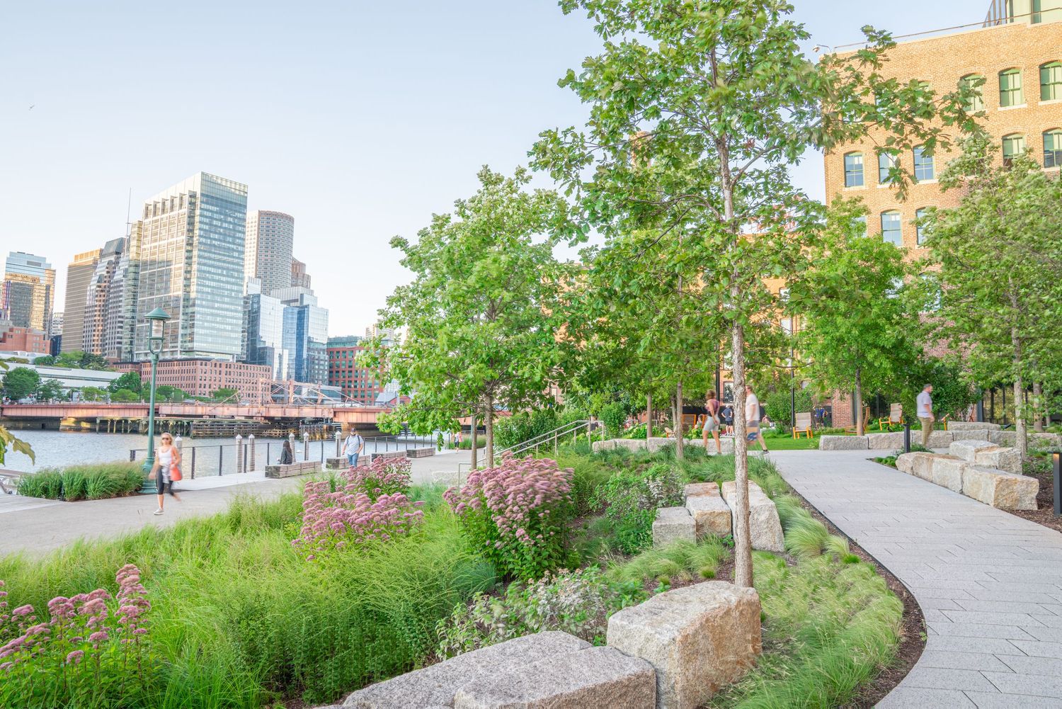 Landscaped waterfront path with flowering plants and modern architecture in the background.