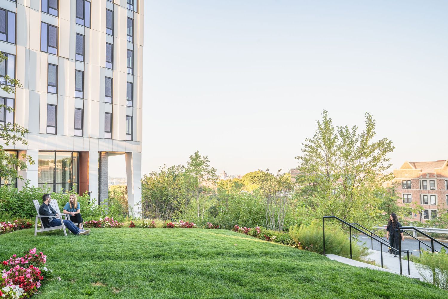 Modern apartment building exterior with landscaped green spaces and concrete walkways leading to the entrance.