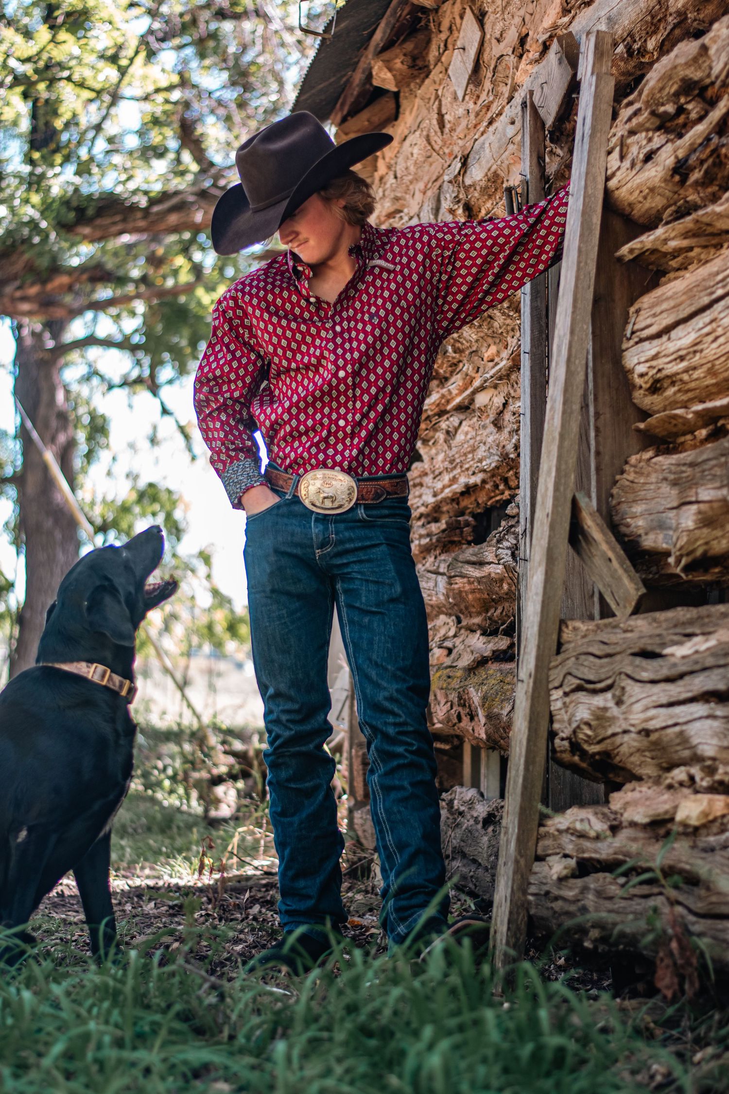 A cowboy in western attire stands near a rustic stone wall with a black dog by their side in a rural outdoor setting.