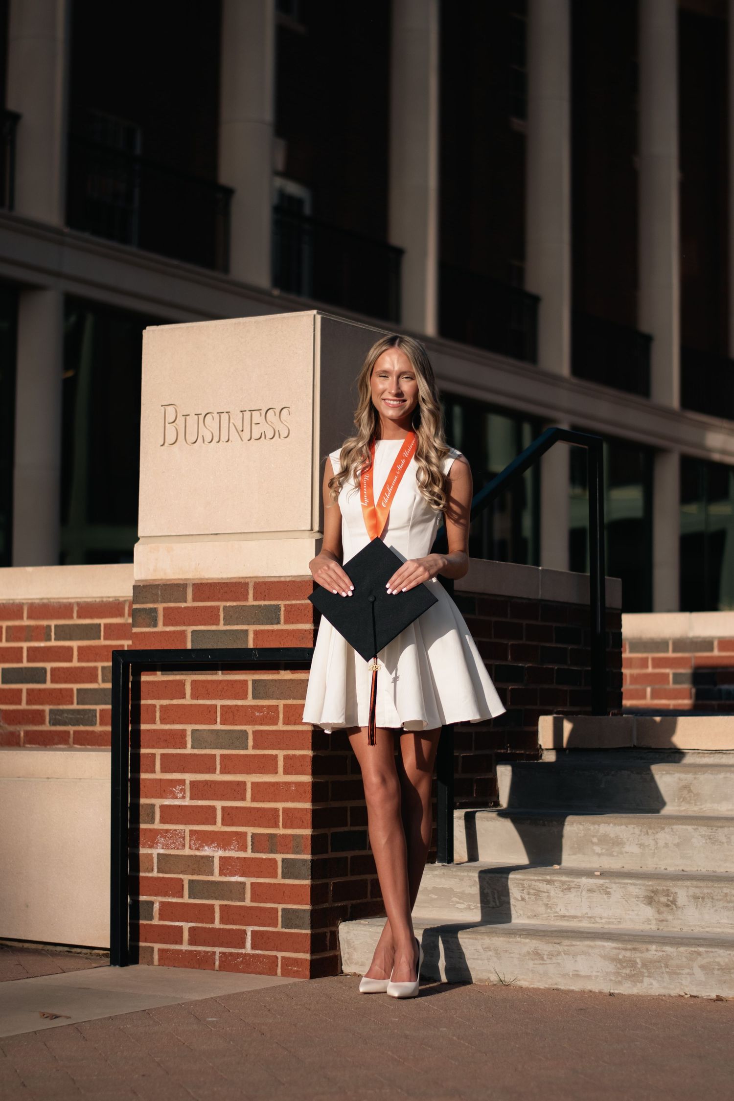 Graduation portrait of a young woman in a white dress with medal and cap, captured at the business school with warm natural light.