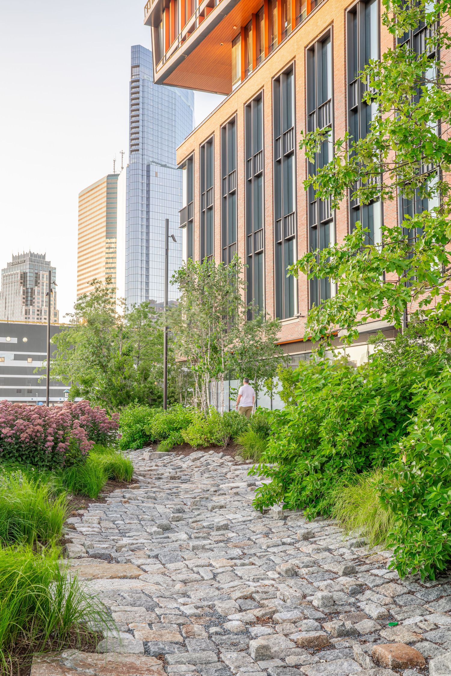 A stone pathway winds through landscaped gardens beside a modern urban building with skyscrapers in the background.