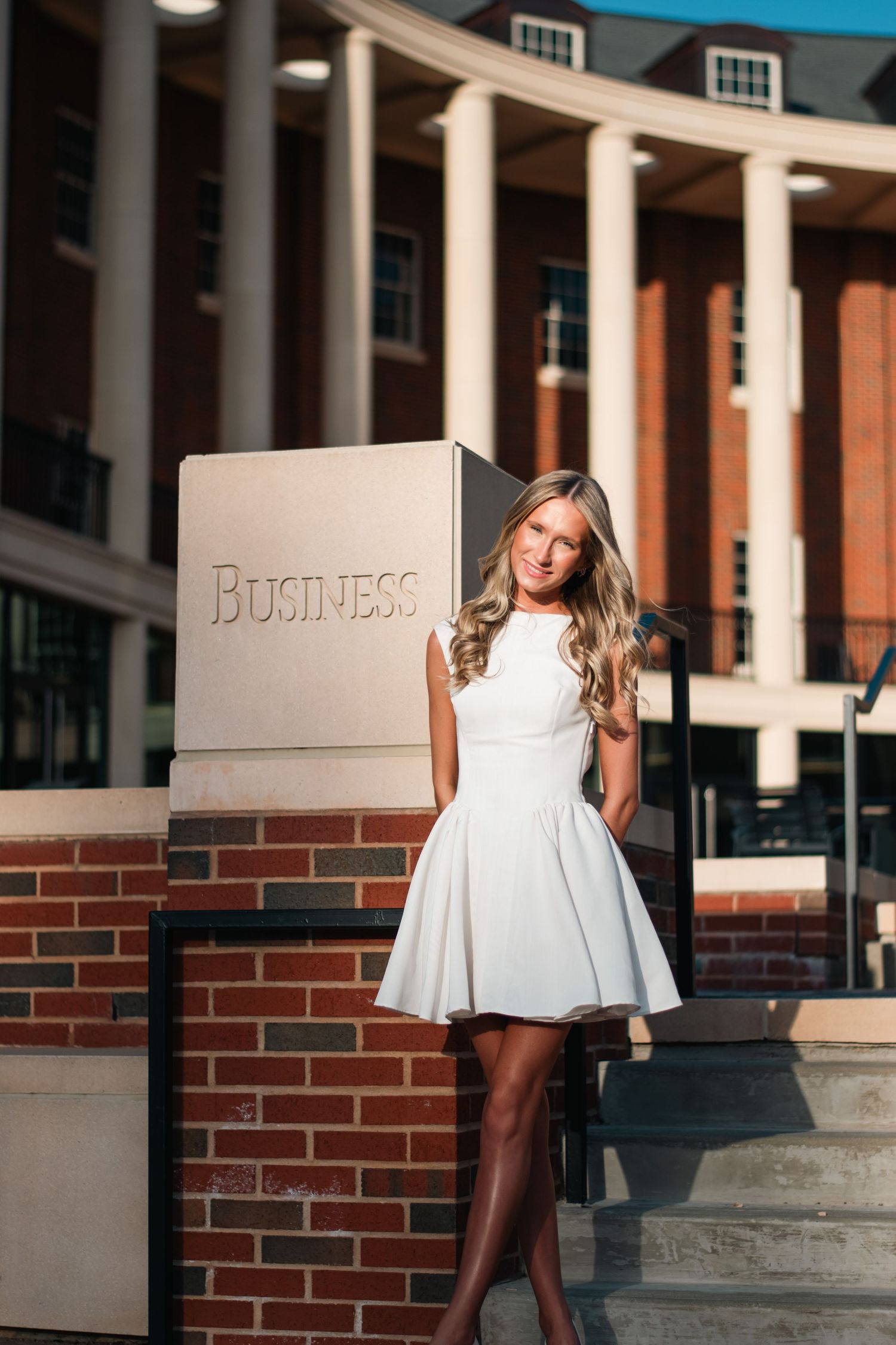 Professional portrait photography of a young woman in a white dress, captured with natural light in front of a classic business school build