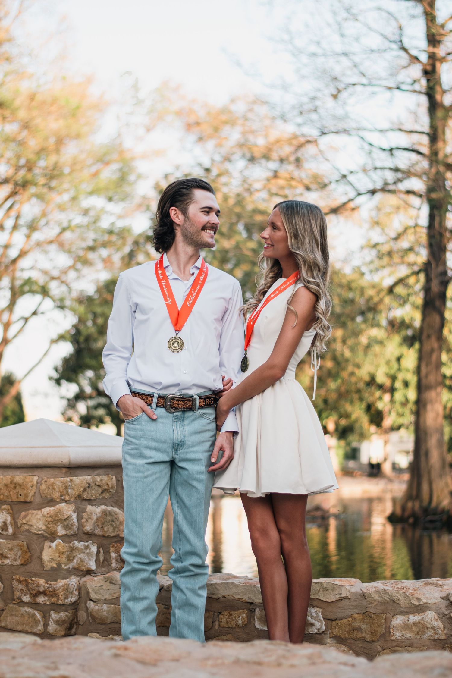 Couple graduation portrait photography with medals, captured outdoors by stonework and water, showcasing joy and achievement in natural ligh