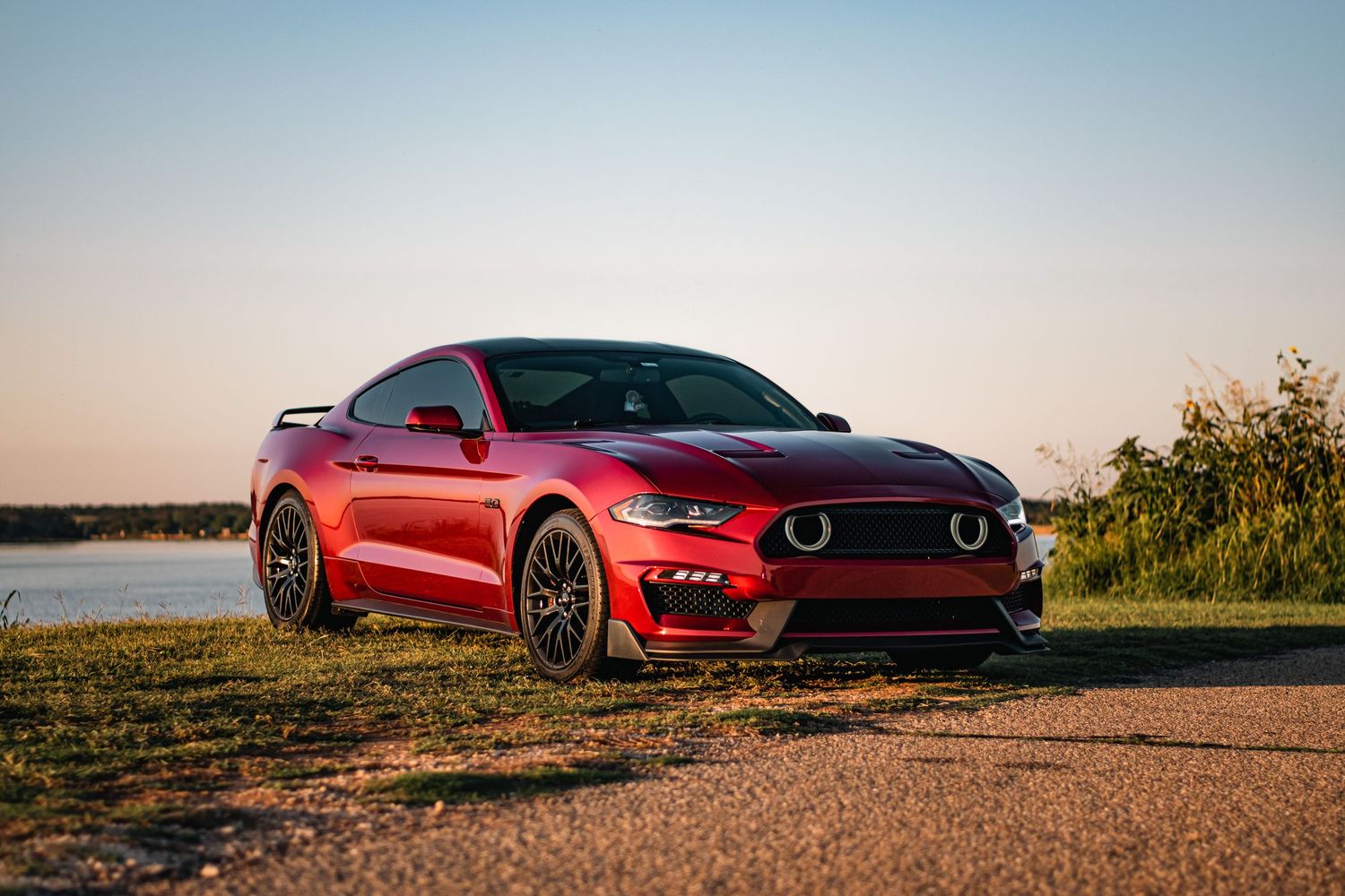 Red Ford Mustang photographed at golden hour by the water, showcasing sleek curves, bold styling, and powerful automotive design.