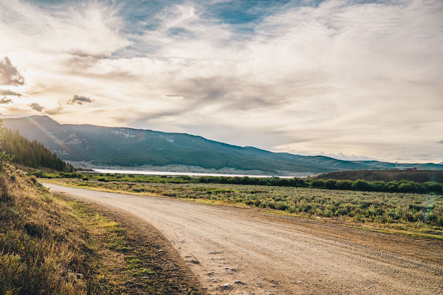A scenic gravel road winds through a vast open landscape with mountains and a lake under a dramatic cloudy sky.