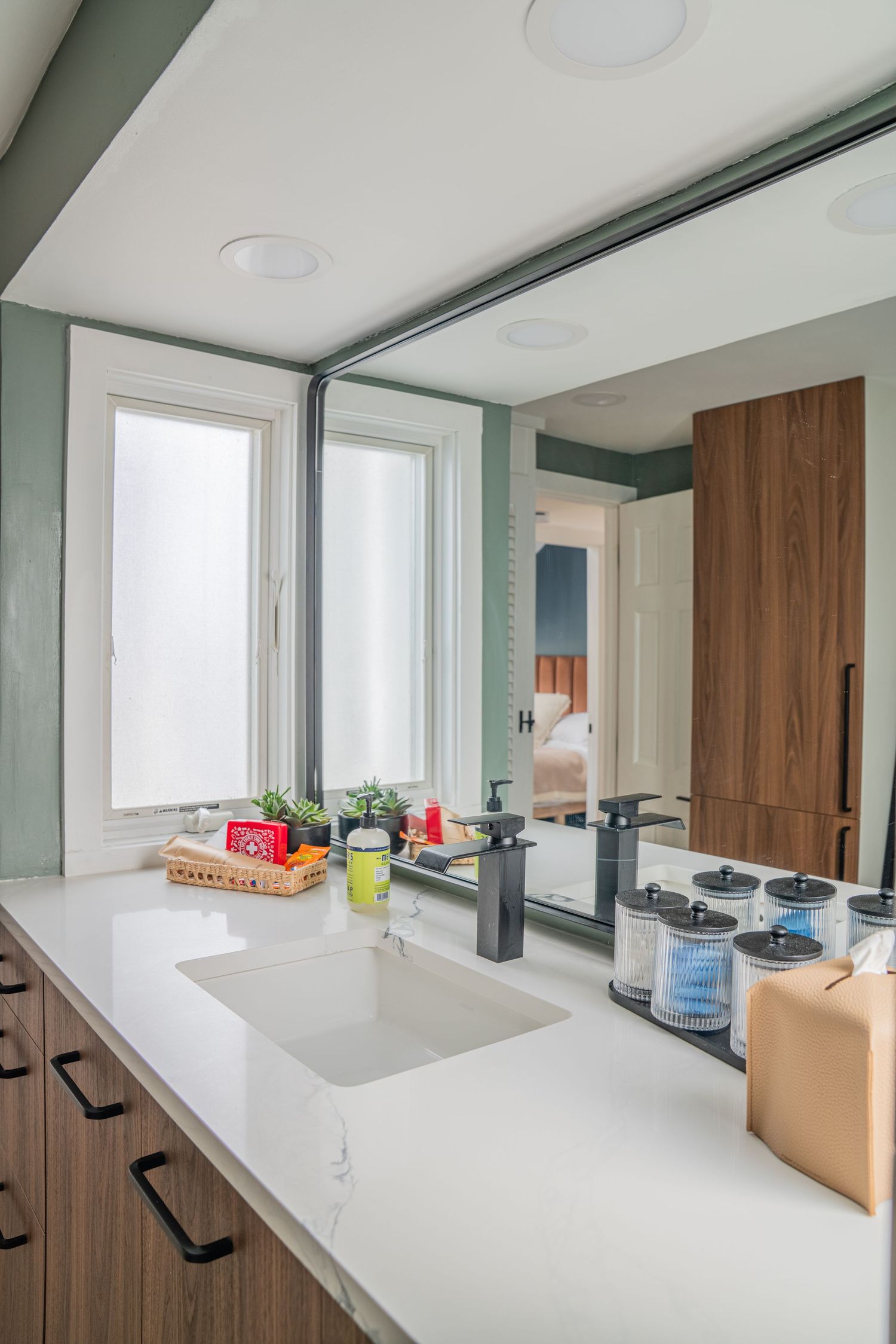 Modern kitchen interior with white countertops and wooden cabinets featuring recessed lighting and large windows.