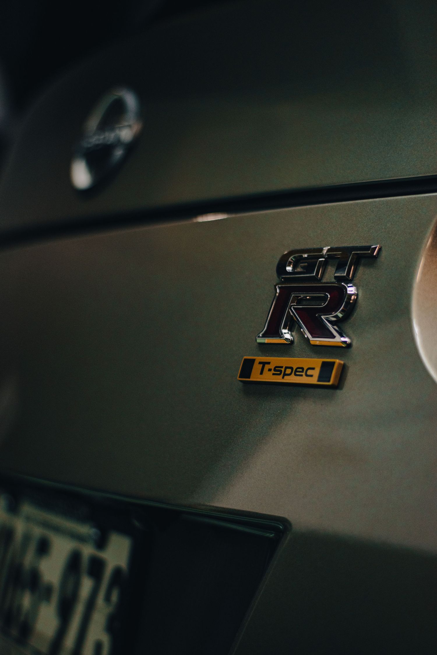 Automotive detail photography of a Nissan GT-R T-spec badge, showcasing sleek design, polished finish, and performance-focused branding.