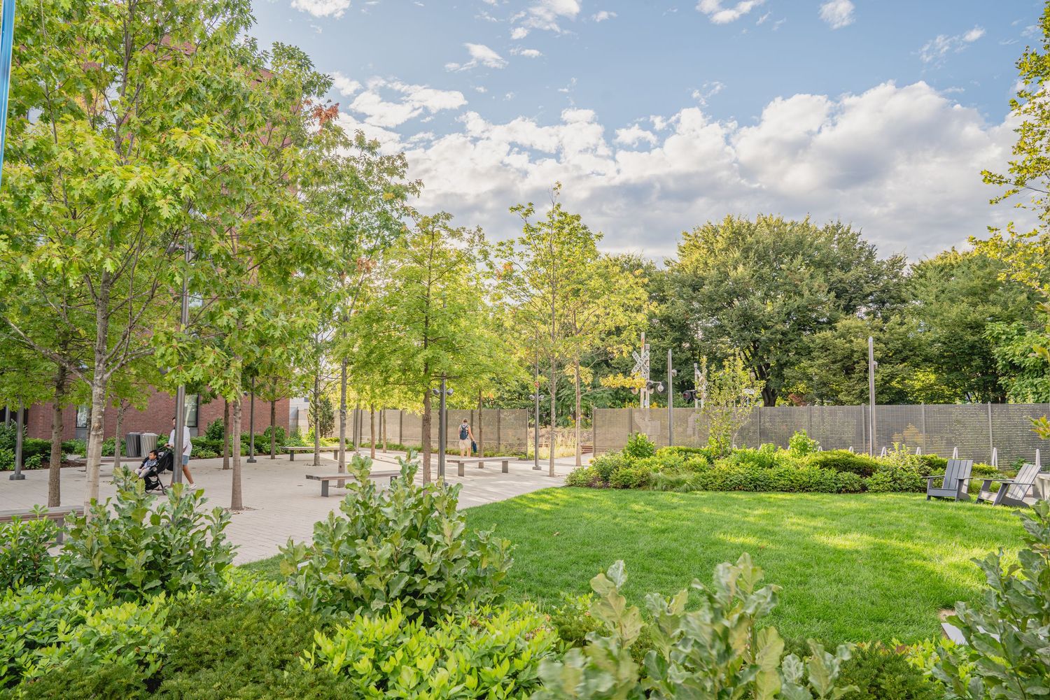Tree-lined pedestrian plaza with brick buildings and landscaped areas under blue skies.