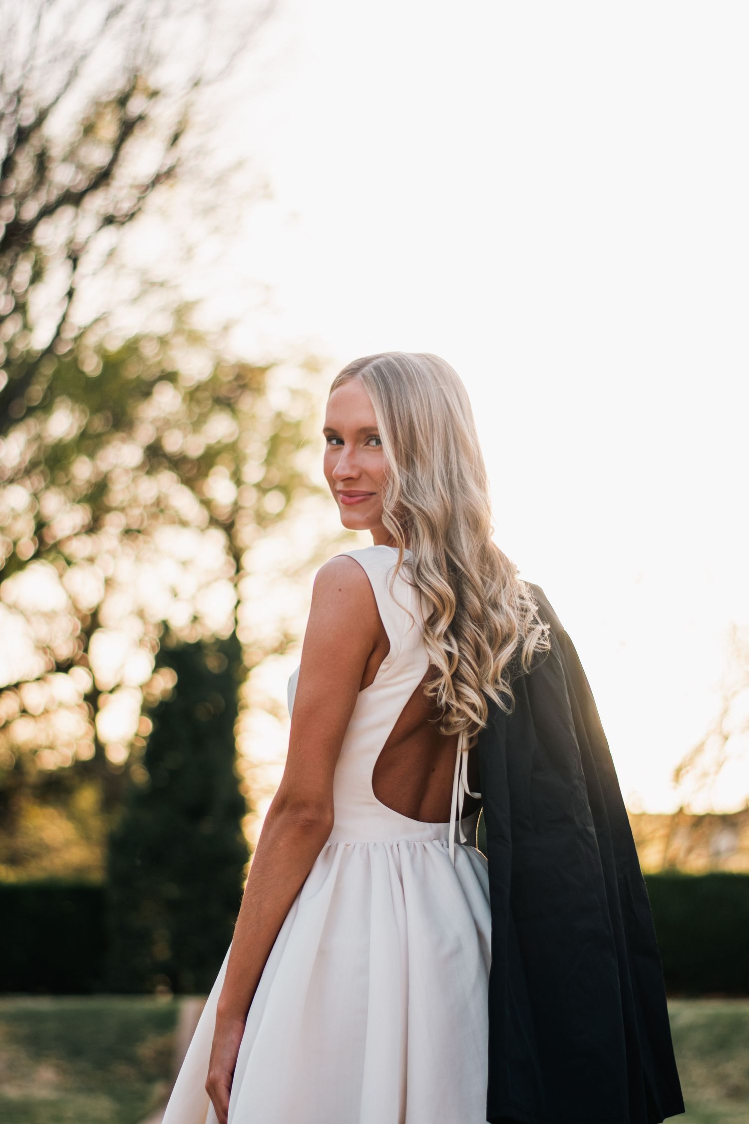 Golden hour graduation portrait of a young woman in a white dress with gown over her shoulder, captured with soft light and natural elegance