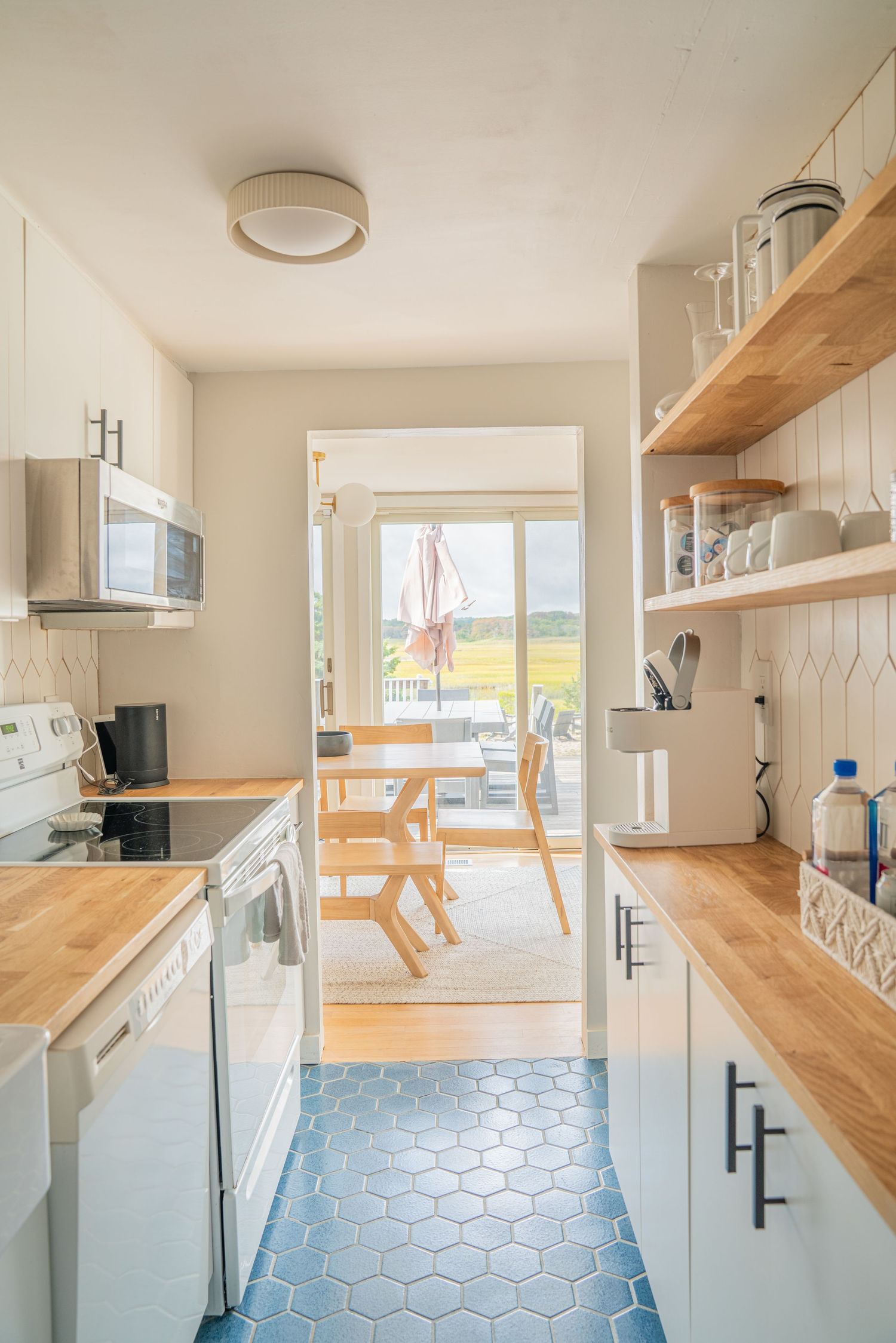 White modern kitchen with wooden open shelving, hexagon tile backsplash, and natural lighting.