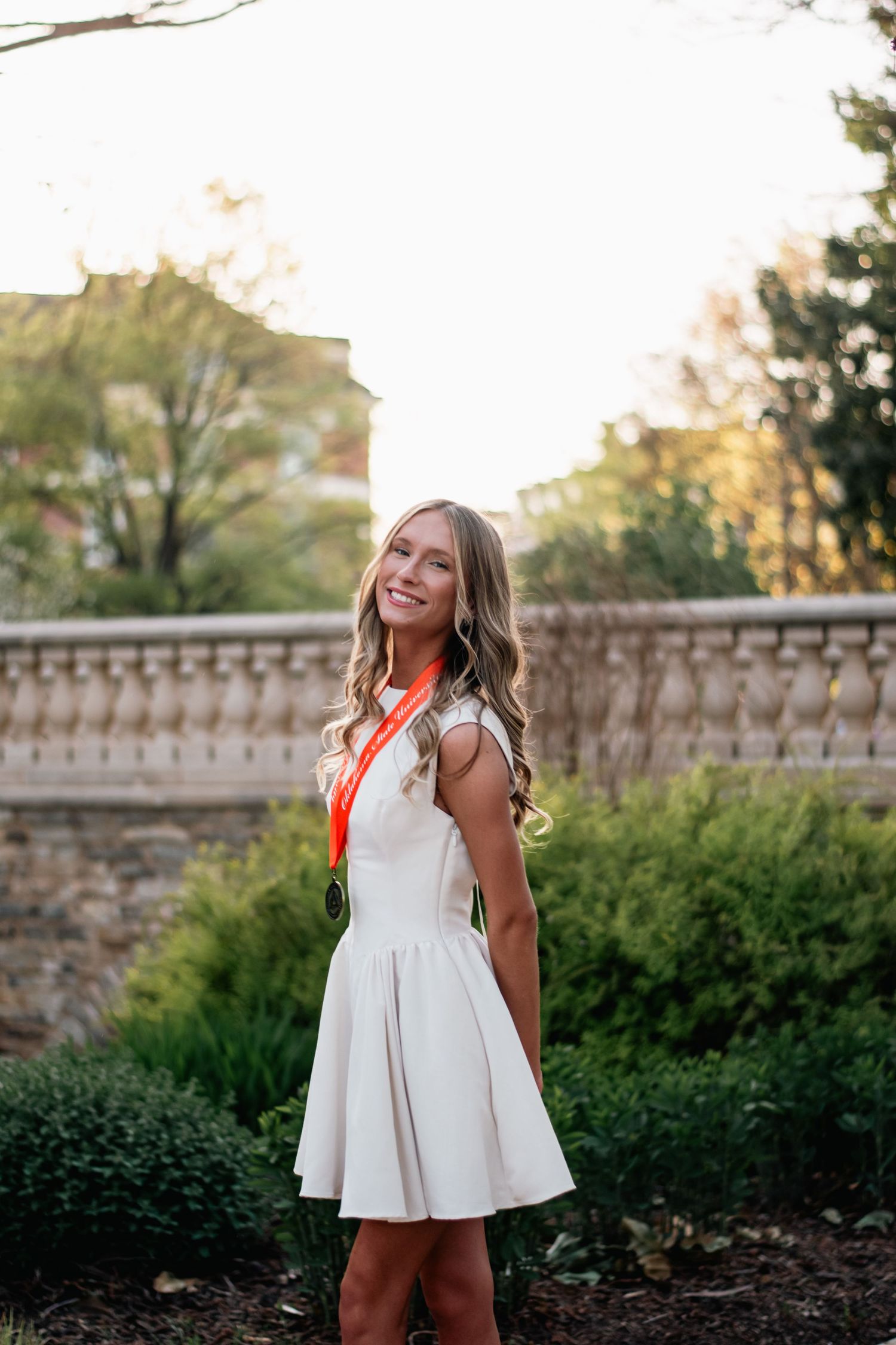 Graduation portrait photography of a young woman in a white dress with medal, captured outdoors in natural light with elegant garden scenery