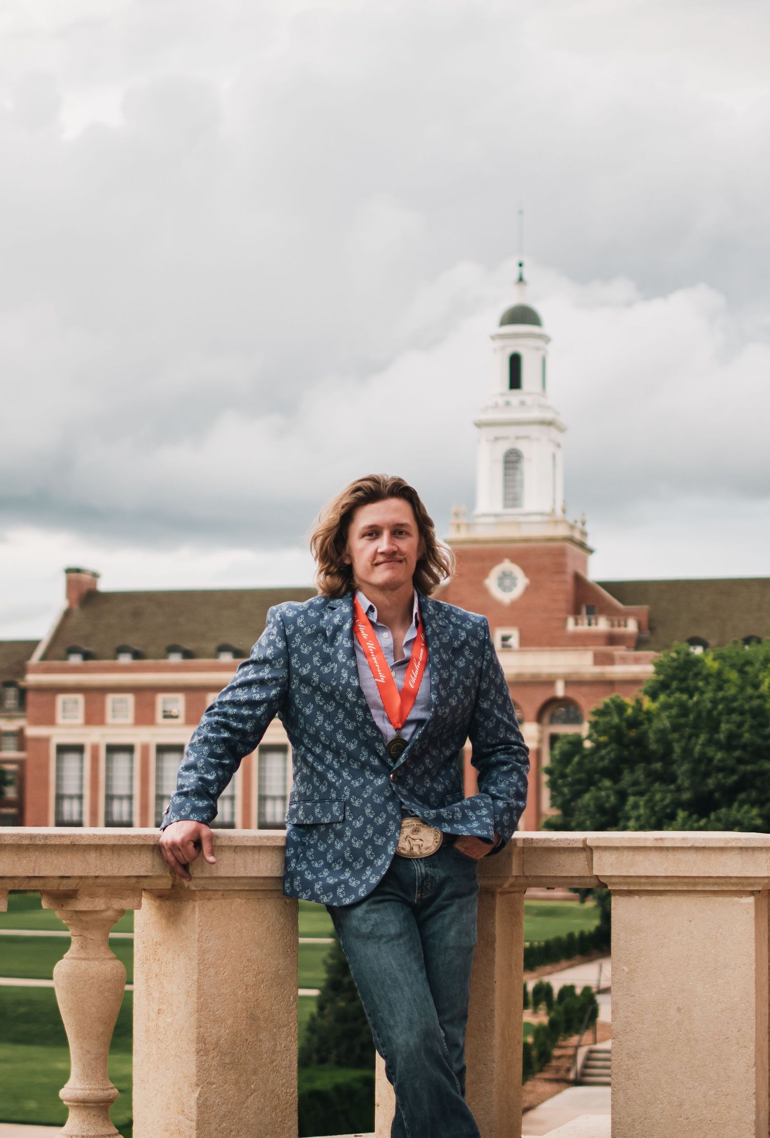 A person in a blue patterned jacket poses playfully on a stone balustrade against a cloudy sky and brick buildings.