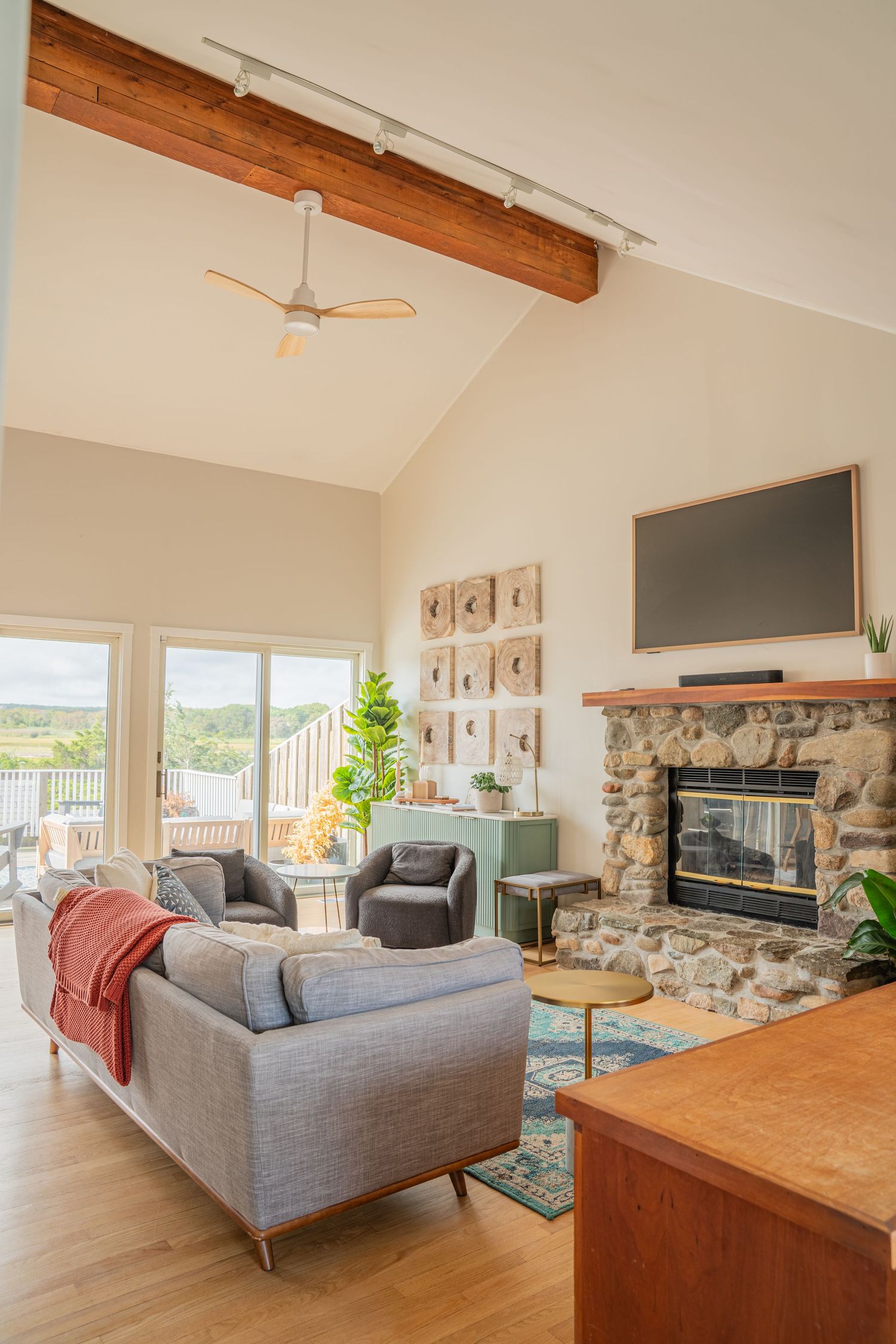 Modern living room features exposed wooden beams, stone fireplace, and gray sectional sofa with panoramic windows.