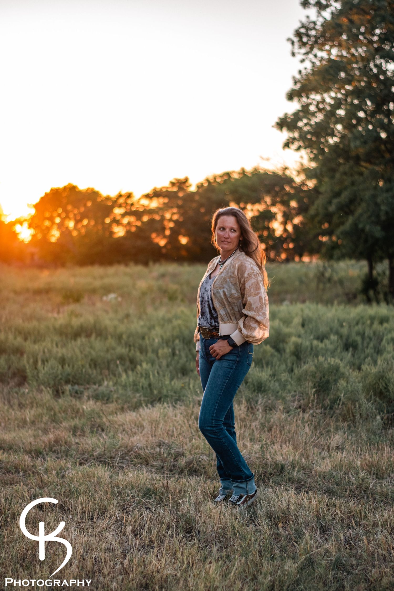 Rustic western portrait photography of a woman in denim and jacket, captured at sunset with warm golden light in an open countryside field.