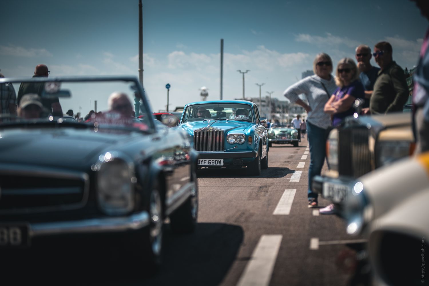 A vintage car show with people admiring classic vehicles lined up on a sunny street, highlighting a bright blue car in the center.