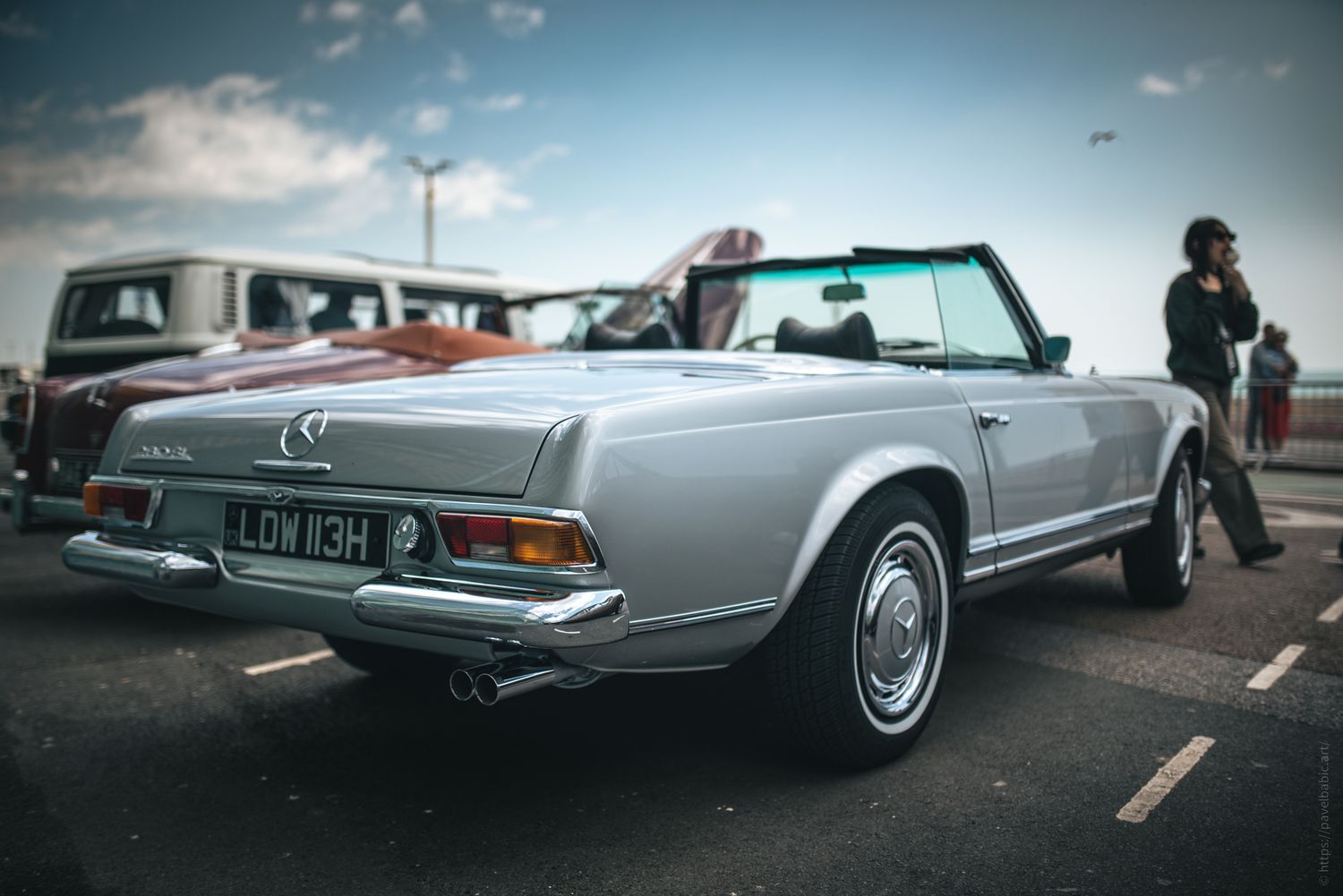 A classic silver convertible car parked, showcasing its sleek design, with a person standing nearby against a cloudy sky backdrop.