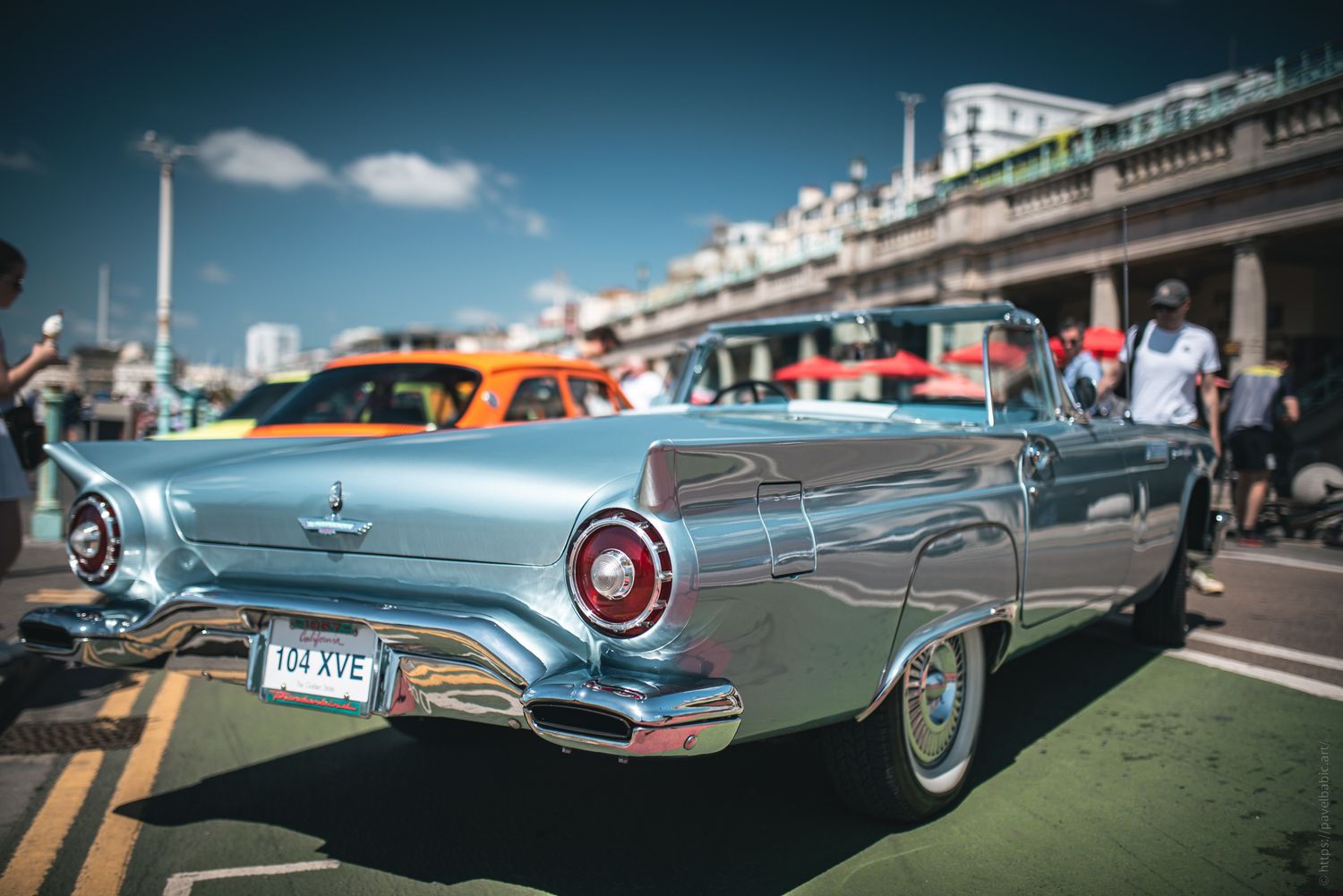 A vintage silver convertible car parked on a sunny street near the beach, surrounded by colorful cars and people.
