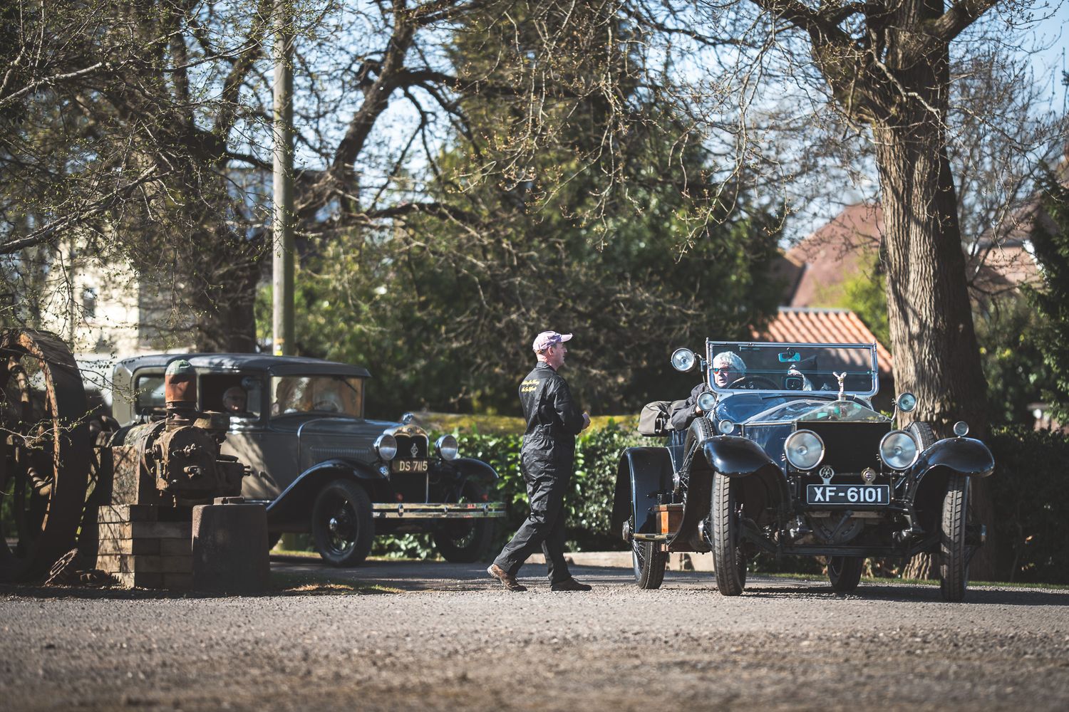 A man walks past vintage cars in a sunny setting, surrounded by trees, suggesting a nostalgic outdoor event.