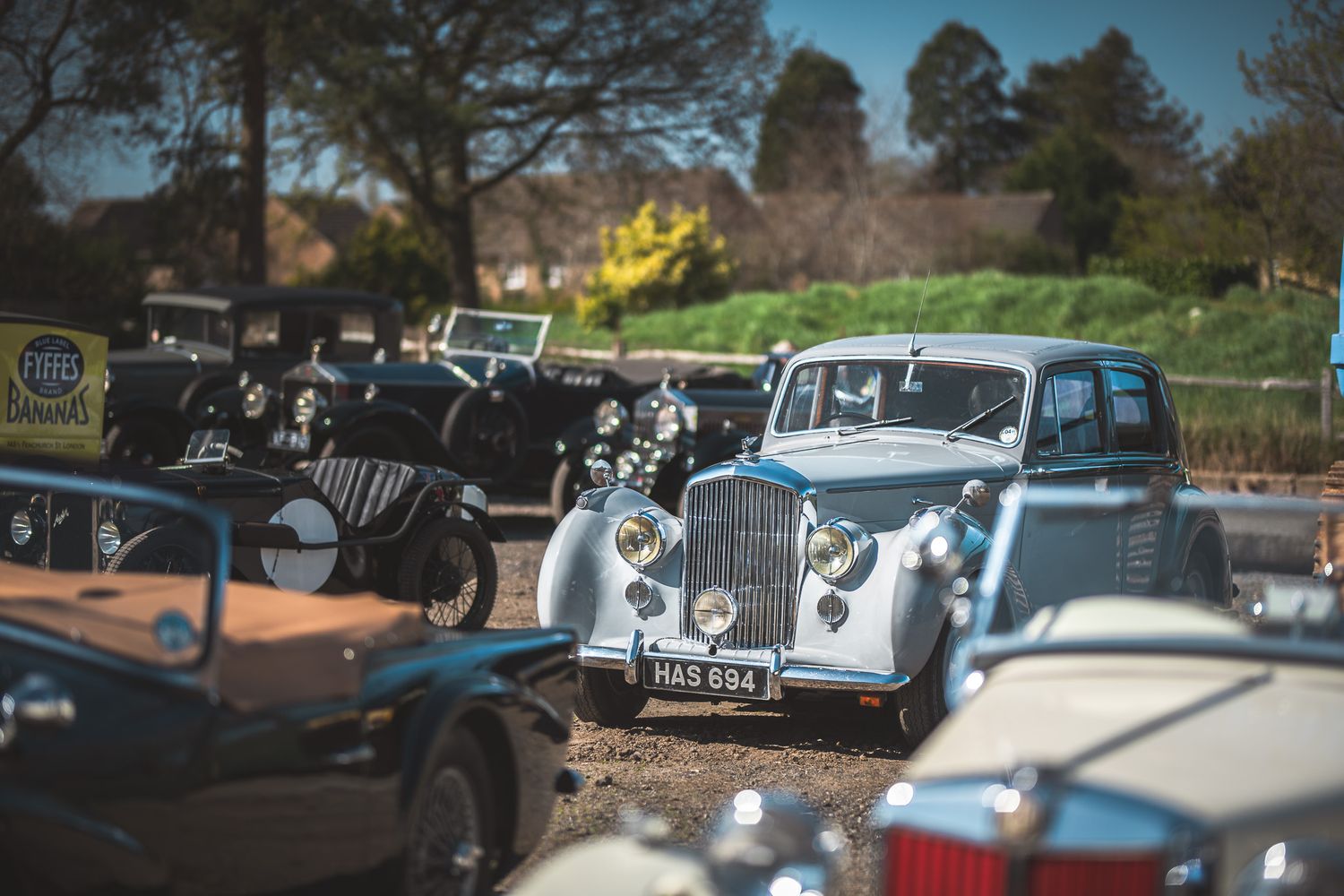 A collection of vintage cars, featuring a silver car in the foreground, parked in a sunny outdoor setting with greenery in the background.