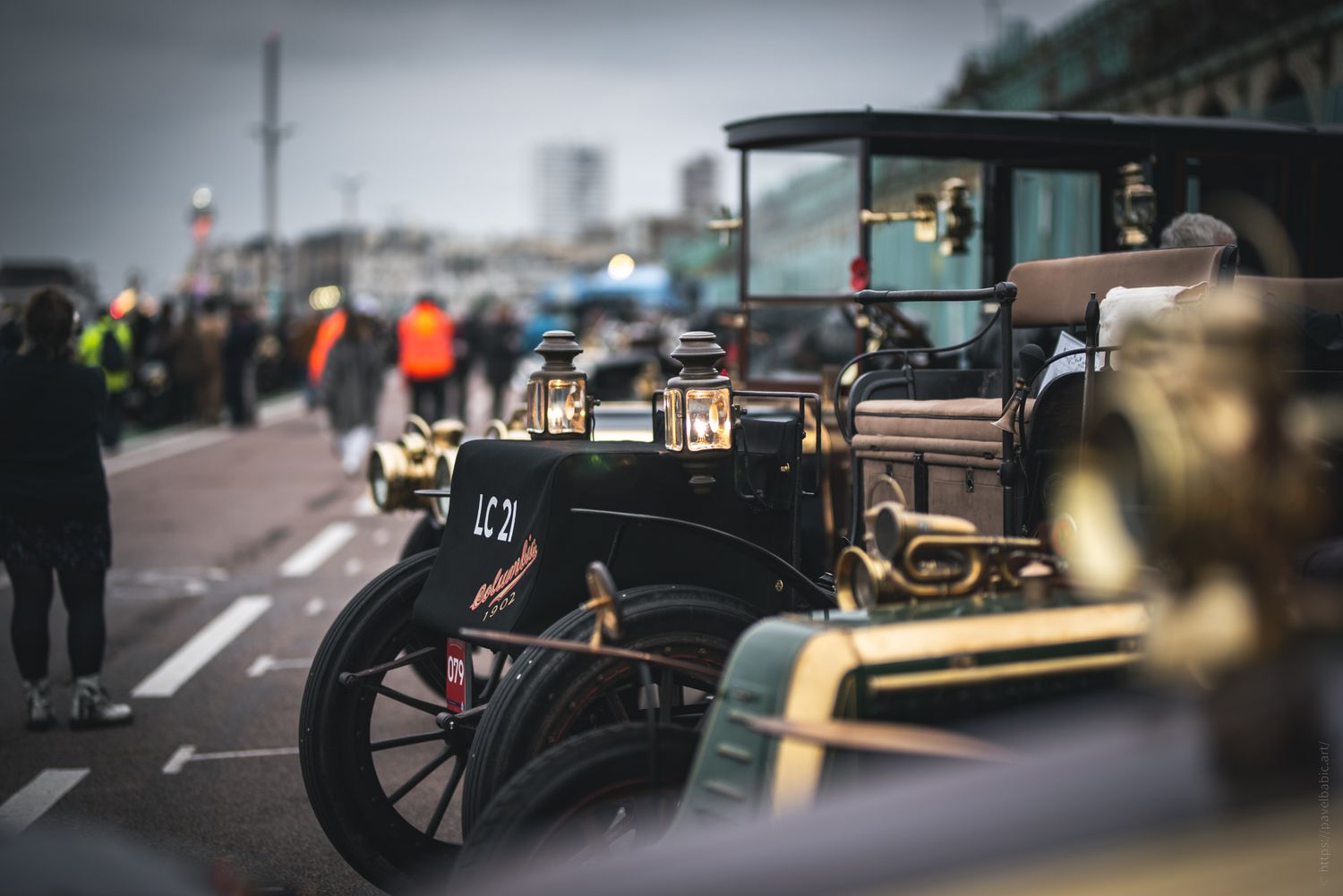 A row of vintage cars lined up along a seaside promenade, with people in bright attire walking nearby on an overcast day.