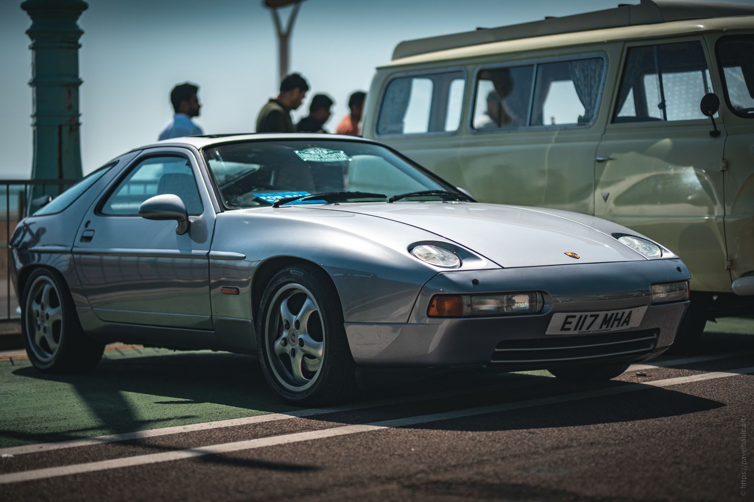 A silver Porsche 928 sports car parked on a sunlit street, with people and a vintage van in the background.