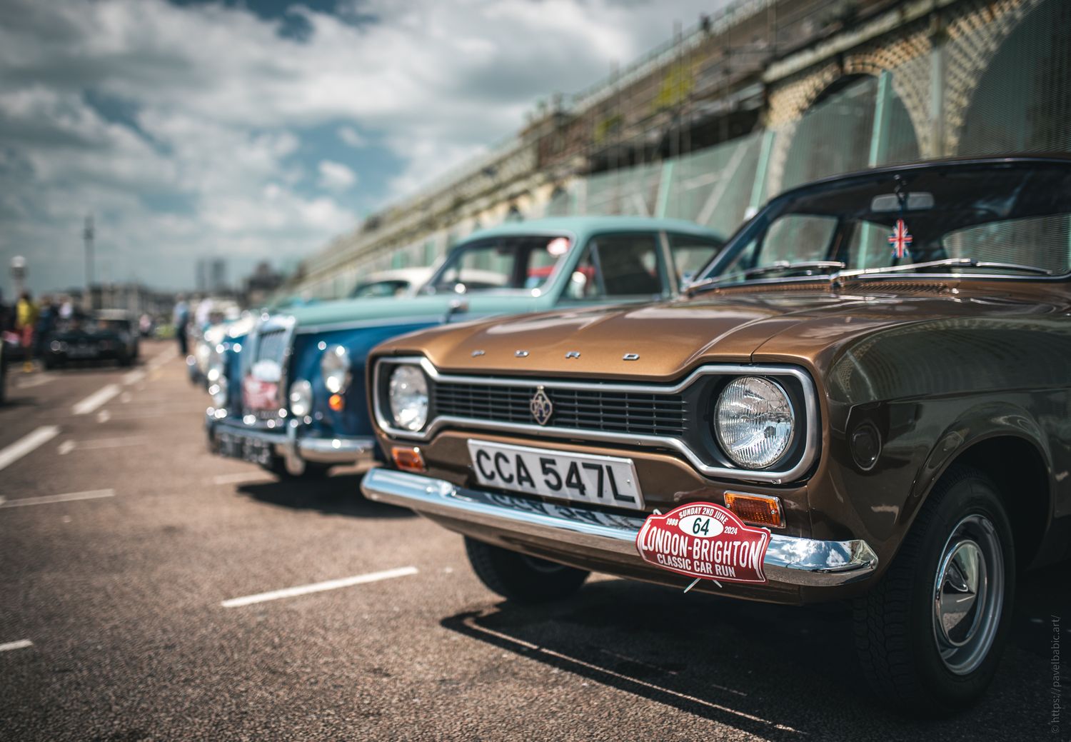 A lineup of classic cars in vibrant colors, including a brown model in the foreground, parked along a sunny promenade.