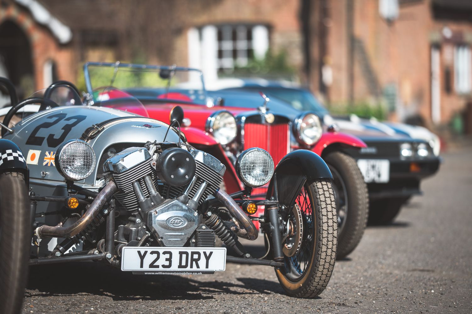 A row of classic cars in various colors, showcasing vintage designs and details, parked outside a rustic building.